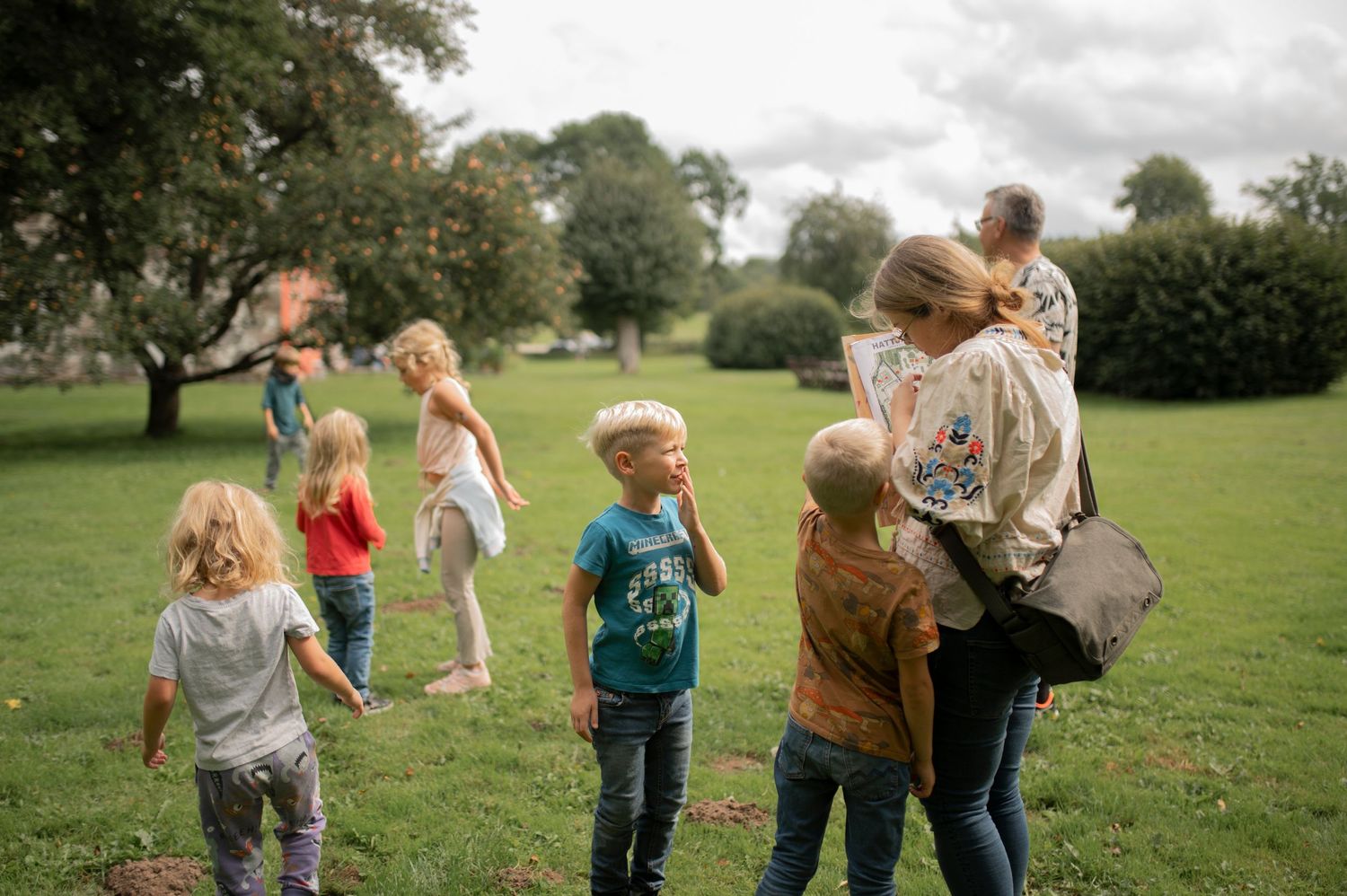 Group gathering in park with children playing on green grass under trees.