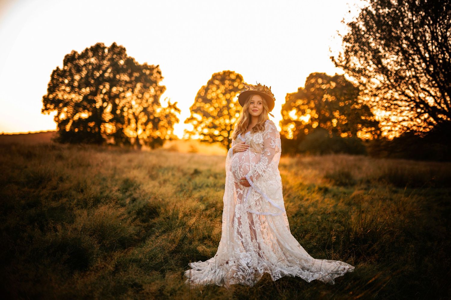Elegant maternity portrait in white lace gown against golden sunset backdrop.