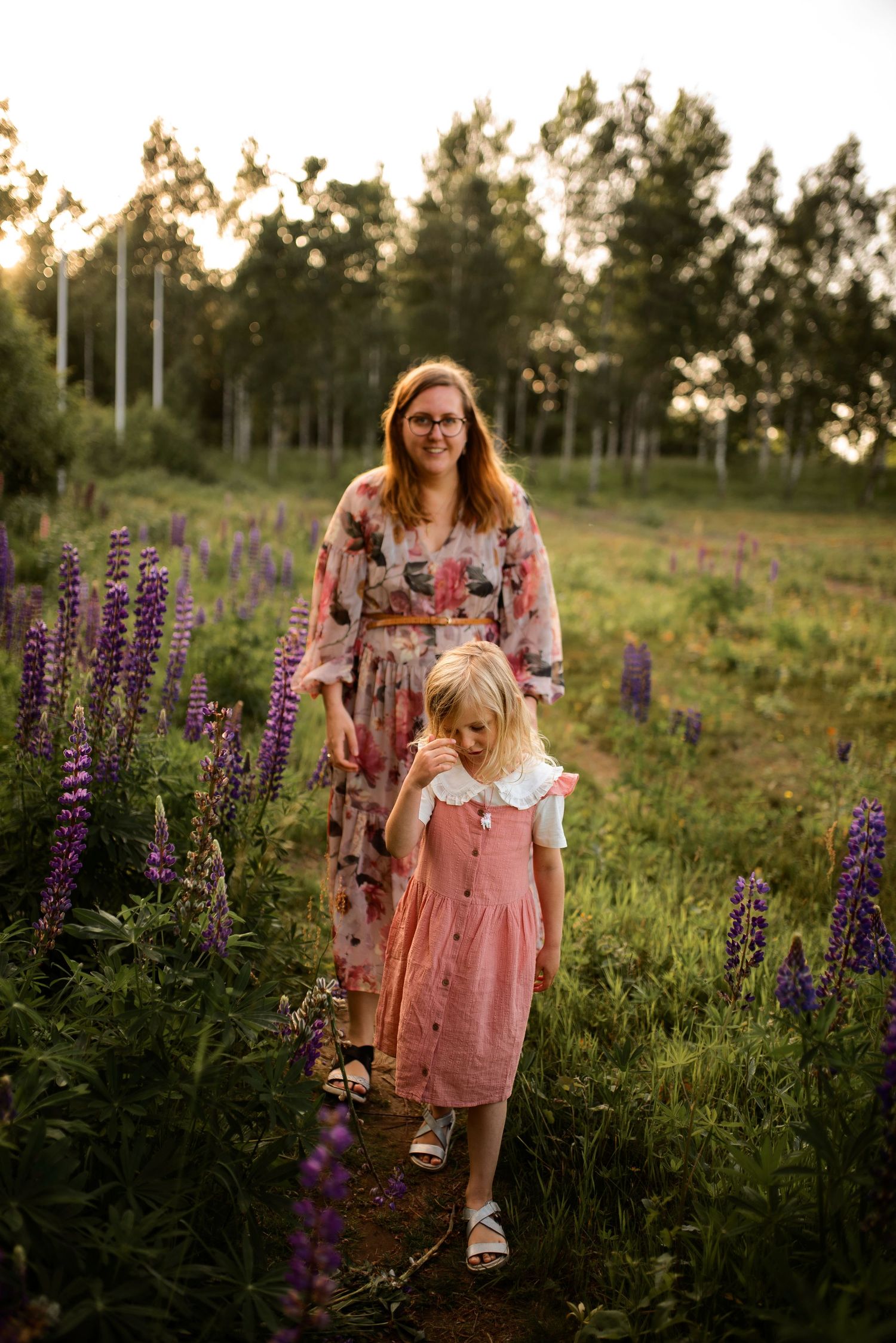 A floral dress and coral pink dress stand among vibrant purple lupine flowers at golden hour.