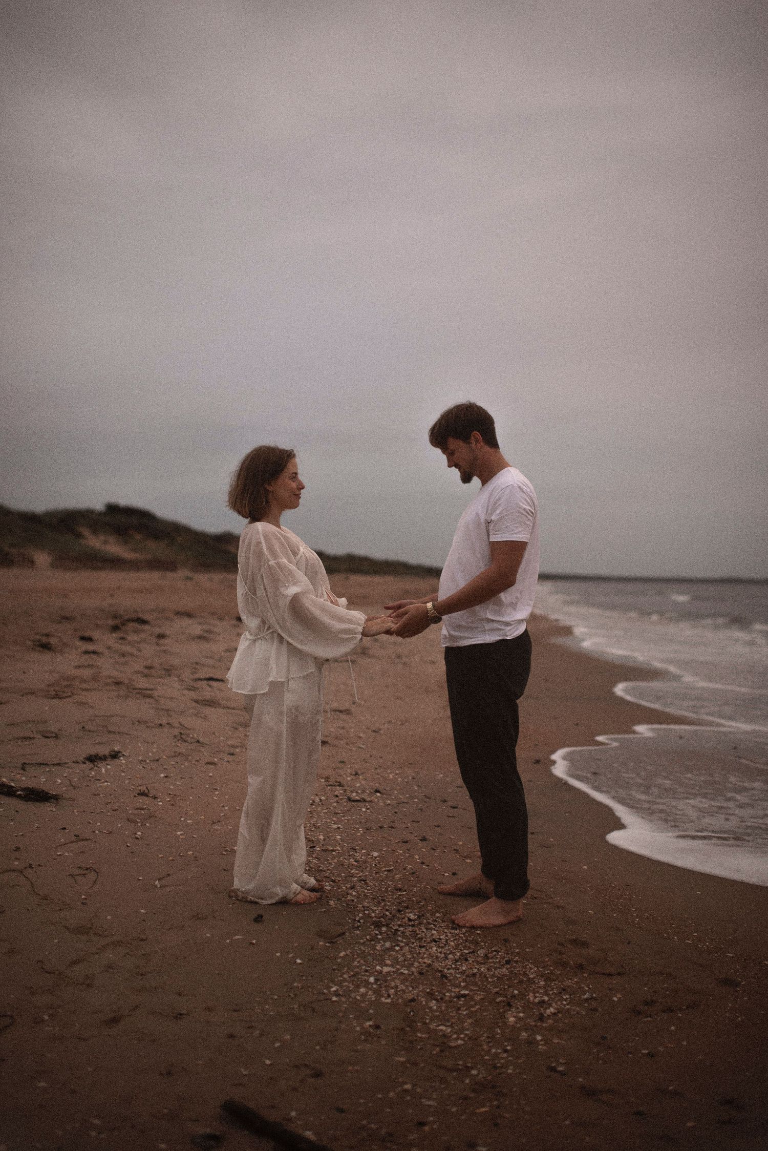 Couple holds hands while standing on beach shoreline during sunset photo session.