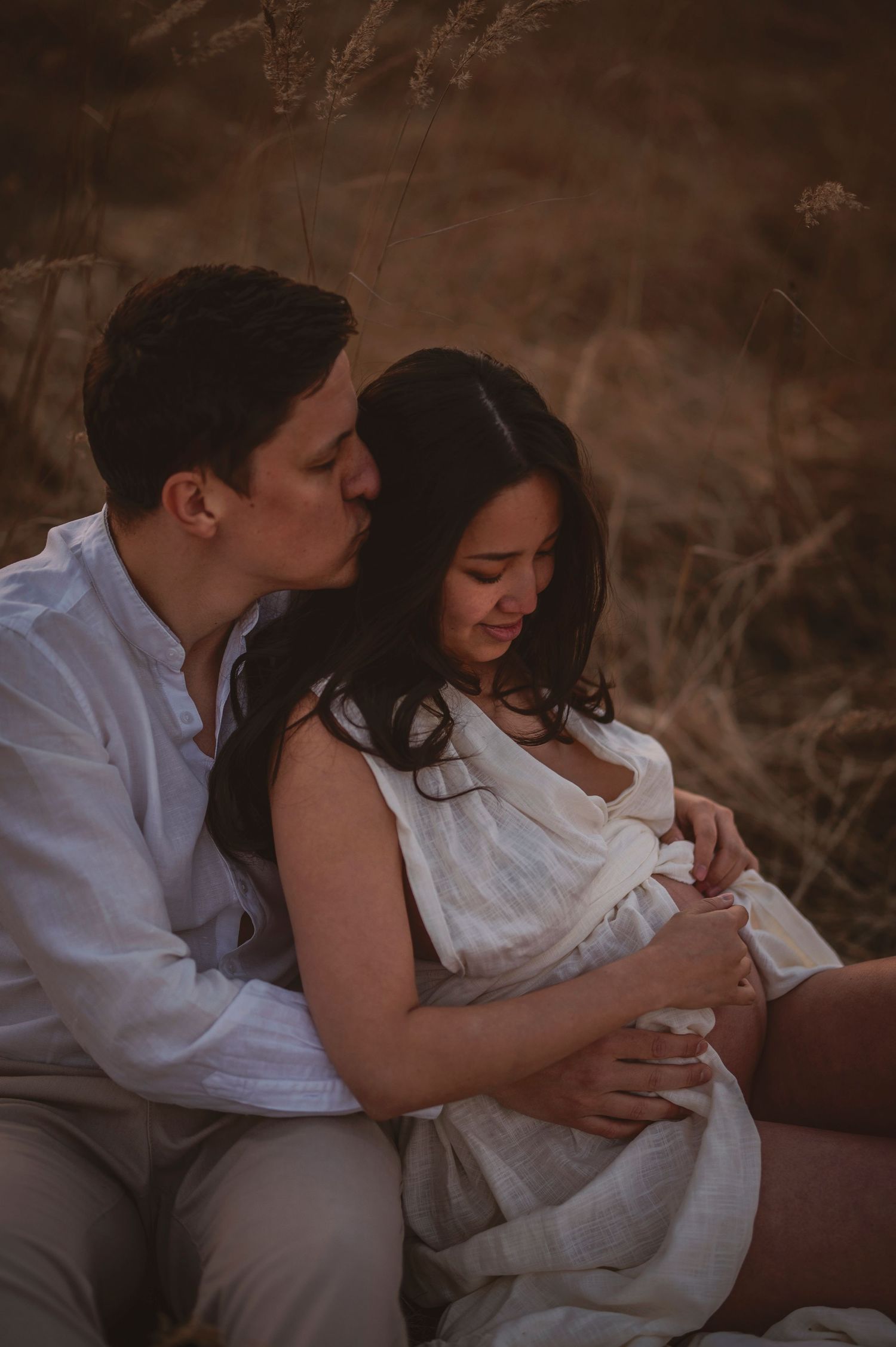 A romantic couple in white clothing share an intimate moment during a rustic outdoor photoshoot at sunset.