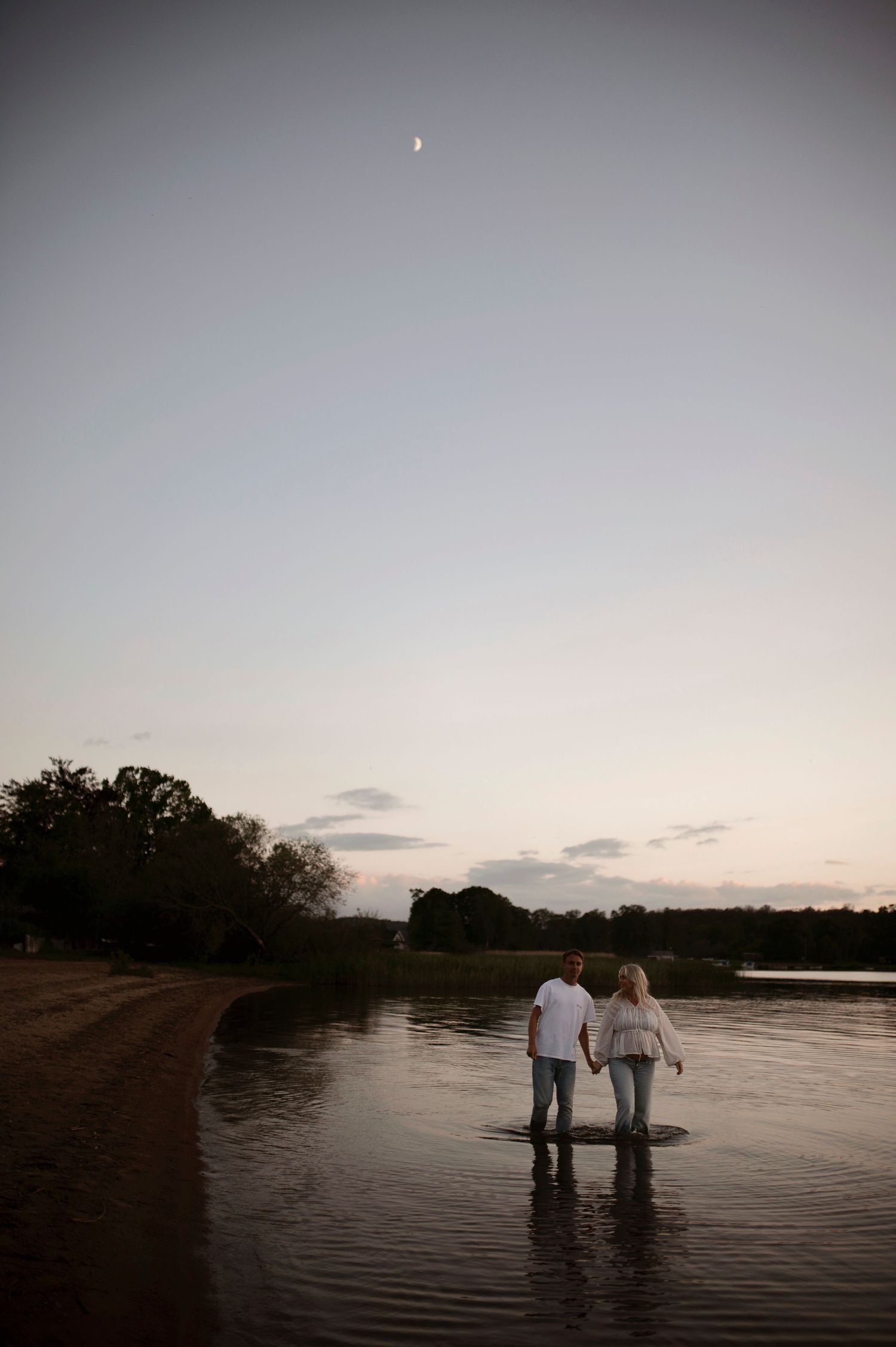A couple stands in shallow water at the edge of a lake during twilight.