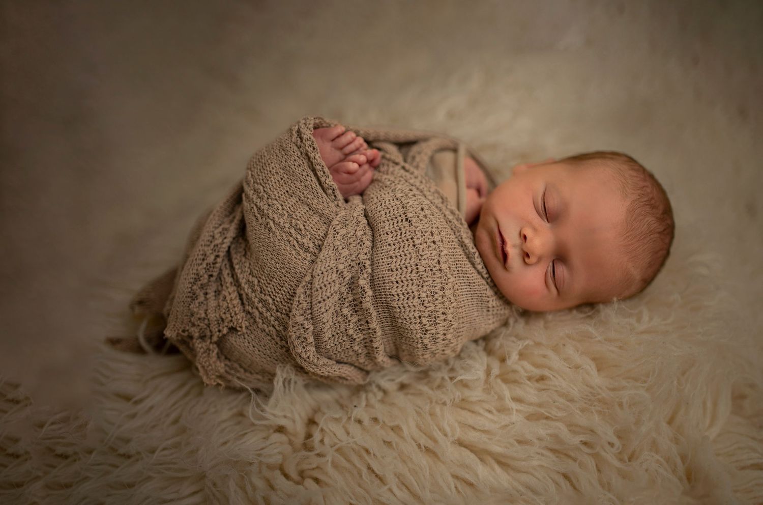 Sleeping newborn wrapped in textured beige sweater on cream fur background.