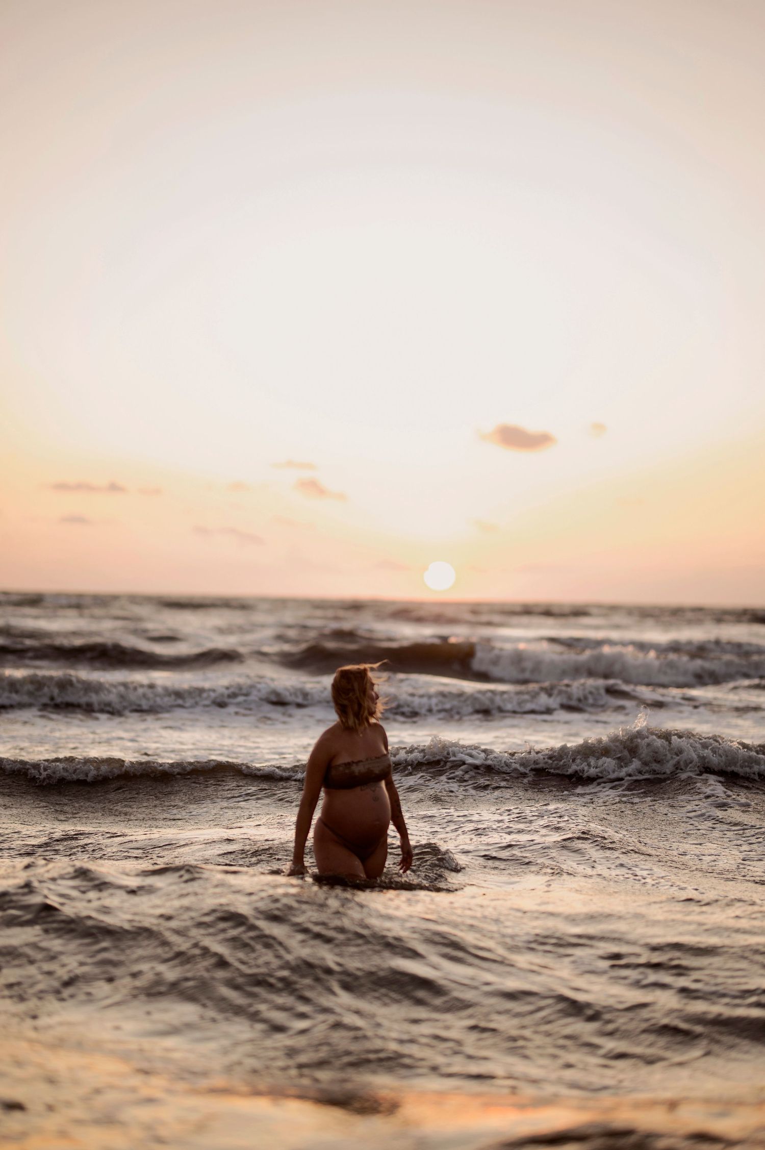 A person sits among crashing ocean waves during a warm sunset at the beach, creating a serene silhouette against the horizon.