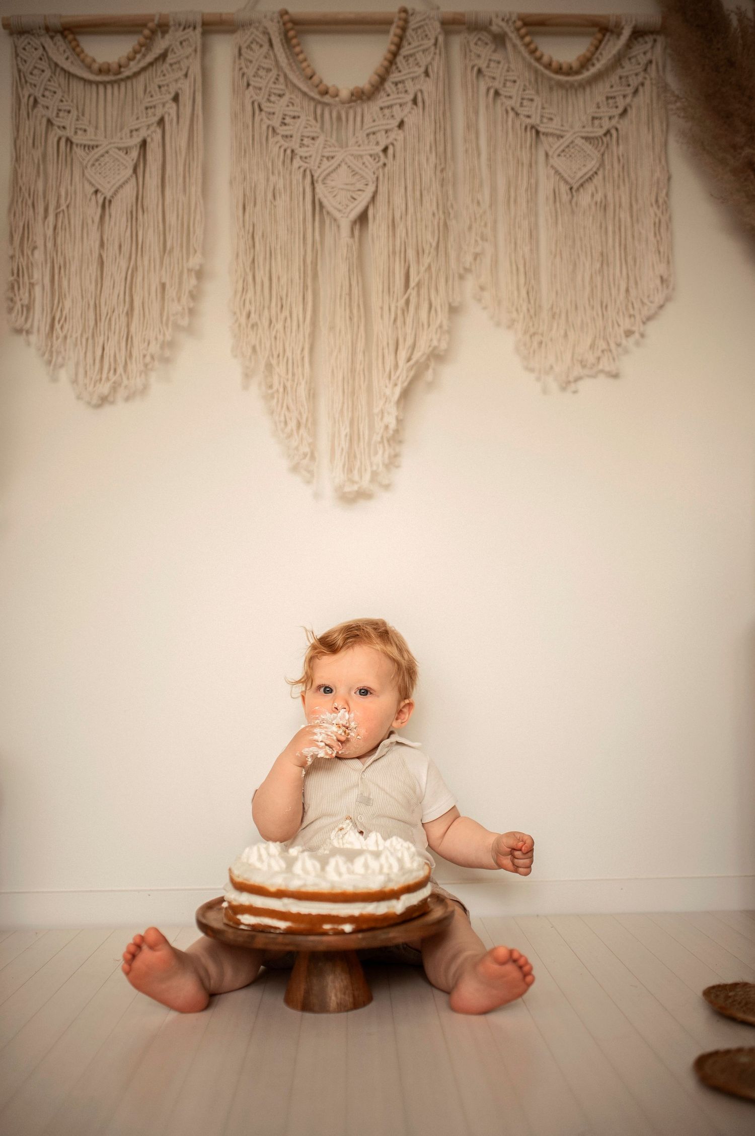 A cake smash photo sequence showing a baby in white sitting next to a decorated cake against a macrame backdrop.