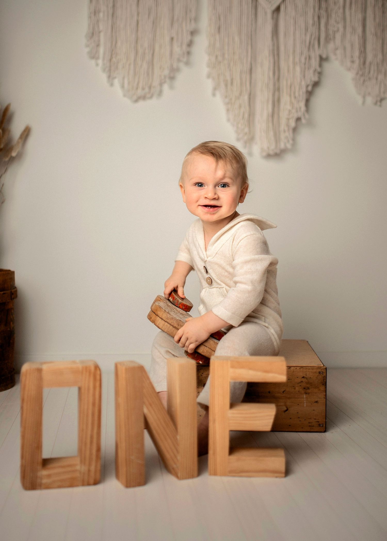 Baby in cream colored outfit sits on wooden box next to large wooden letters spelling ONE against neutral backdrop.