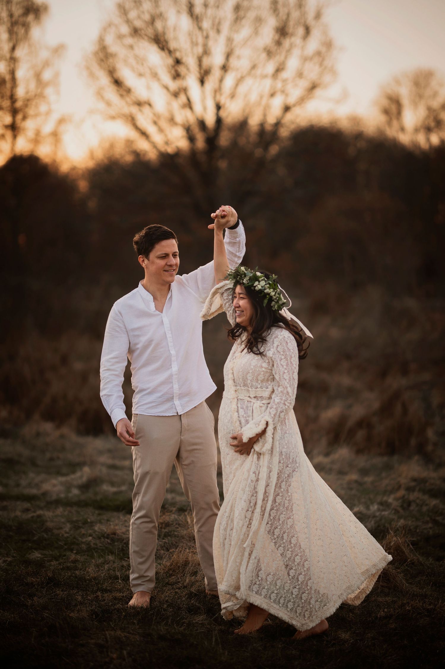A couple shares a romantic moment at sunset, with the person in white wearing a flowing lace dress and floral crown.