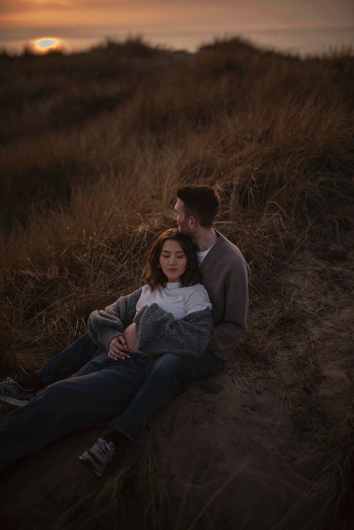 A couple embraces on a hillside against a moody sunset backdrop of dry grass and fading light.