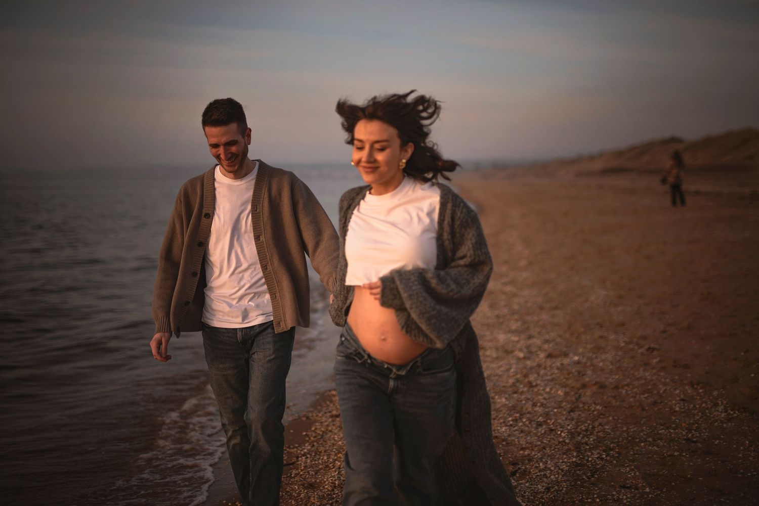 A couple strolls together on a sandy beach during golden hour wearing casual clothing.