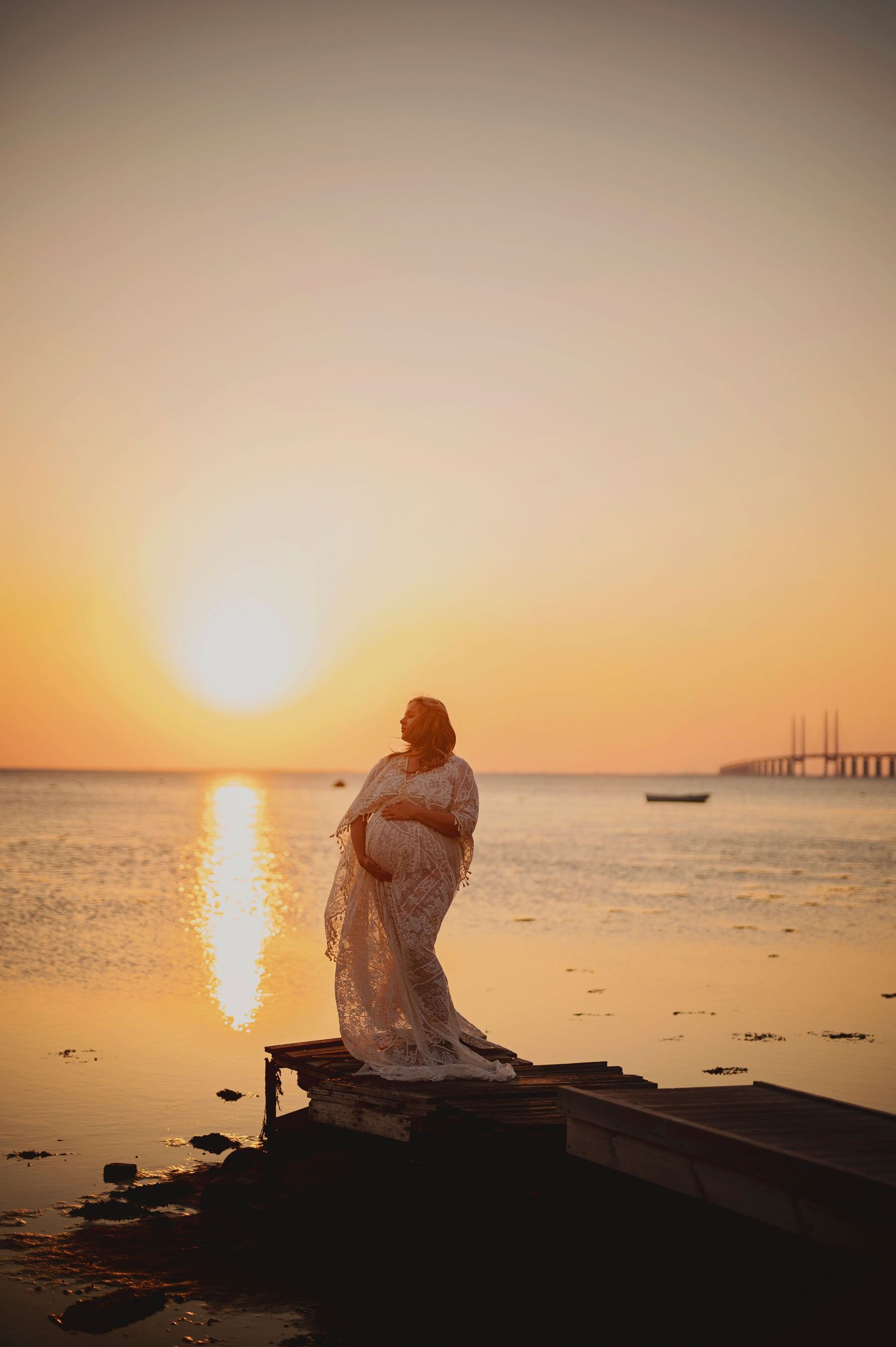 A silhouette stands on a boat during golden sunset over calm waters with a bridge in distance.