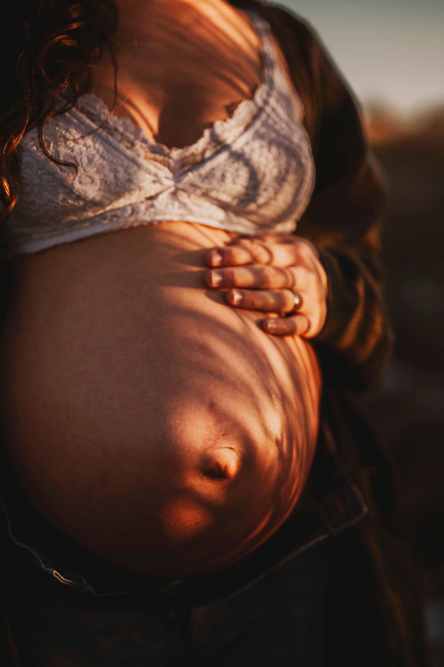 Close-up of pregnant belly with hands cradling bump, wearing lace bra in warm sunset light.