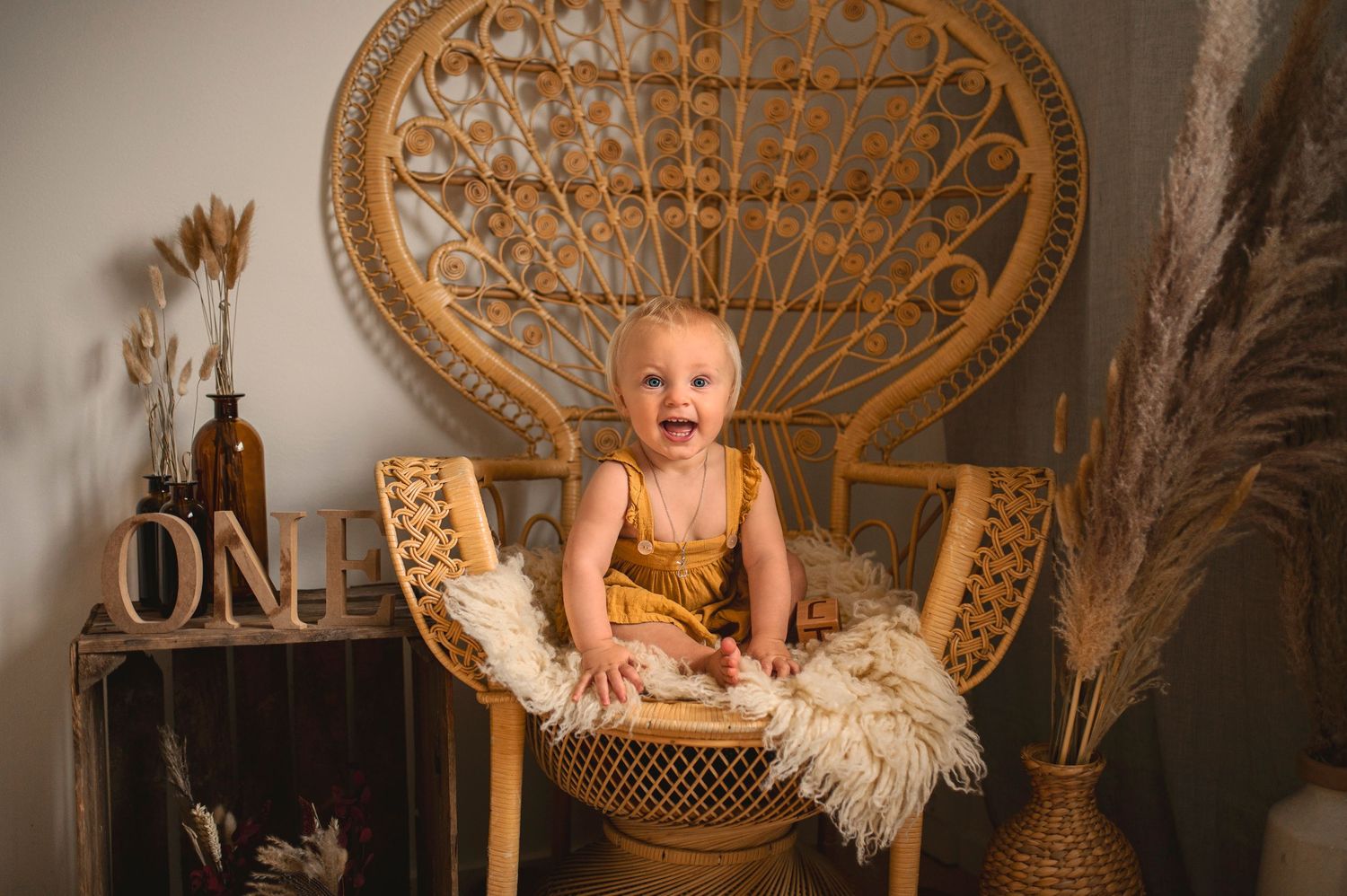 Baby in yellow outfit sits on peacock chair surrounded by boho decor and ONE sign.
