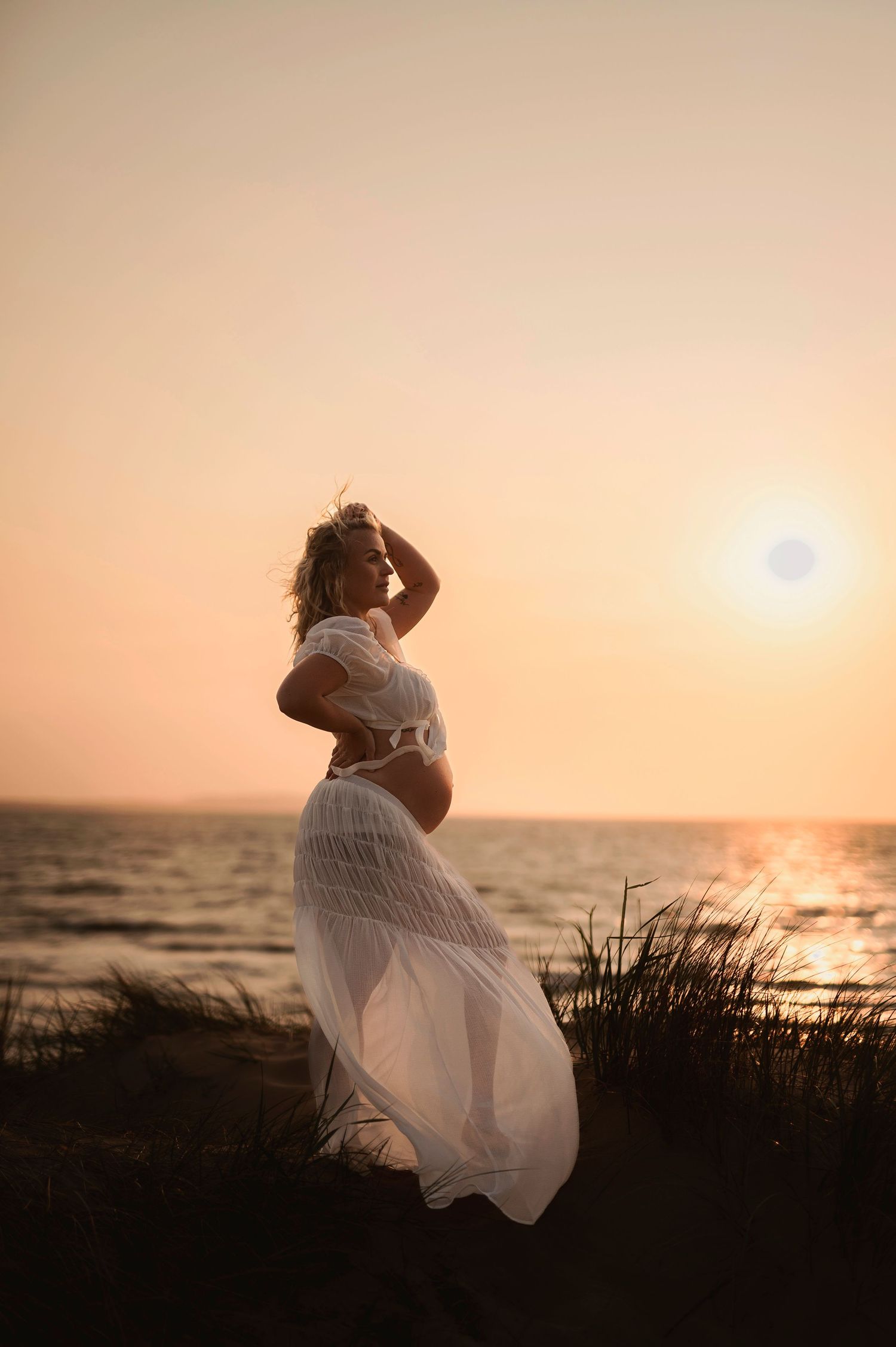 Silhouette of figure in flowing white dress against sunset ocean backdrop.