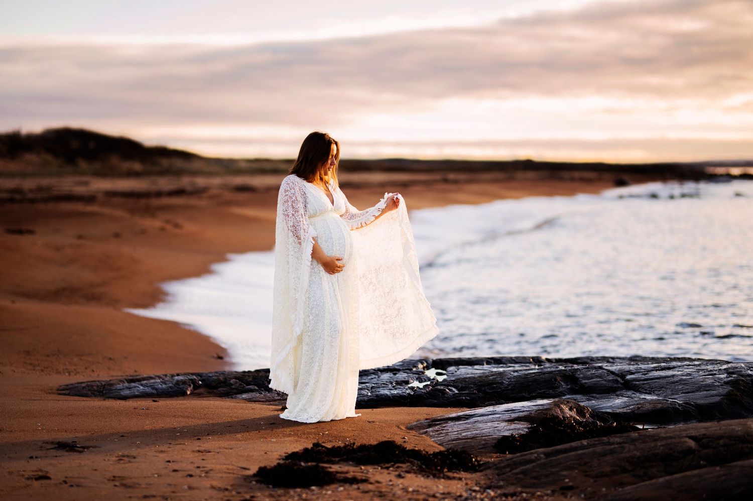 Flowing white dress catches sunset light as figure stands on rocky beach near ocean waves.