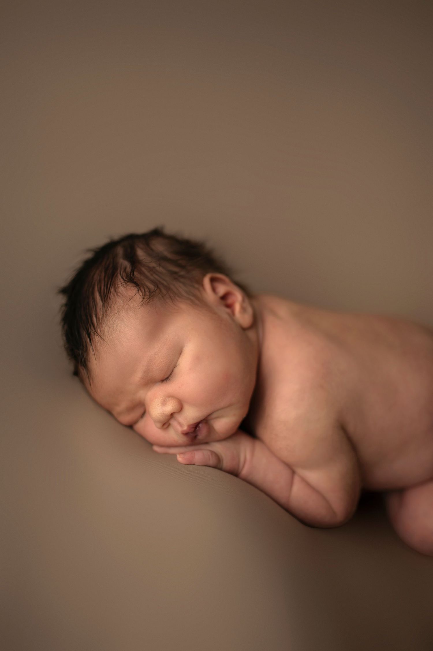 Side profile of sleeping newborn against neutral brown background.