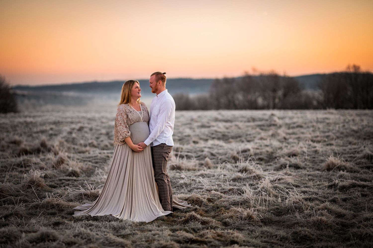 Sunset maternity portrait of a couple in neutral tones standing in a frosty field.
