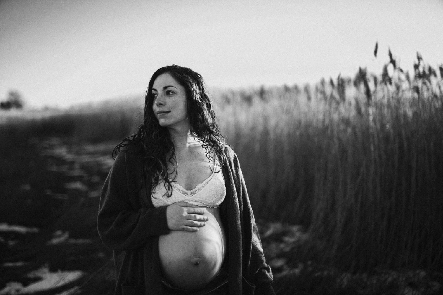 Expectant mother in black coat poses in front of tall reeds during a maternity photoshoot.