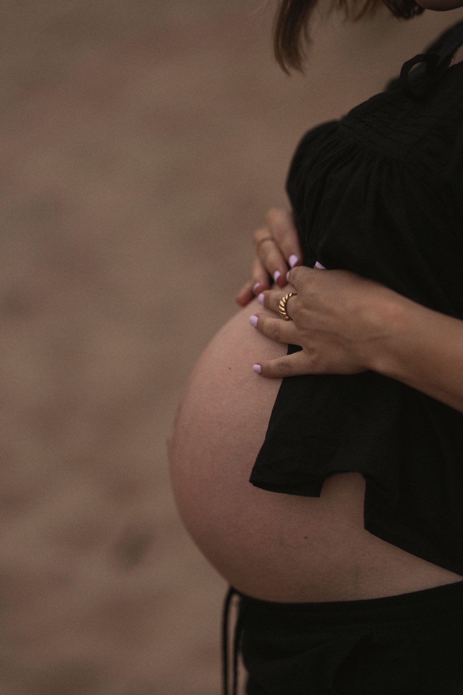Close-up of a pregnant belly with hands wearing rings resting on it against a dark background.