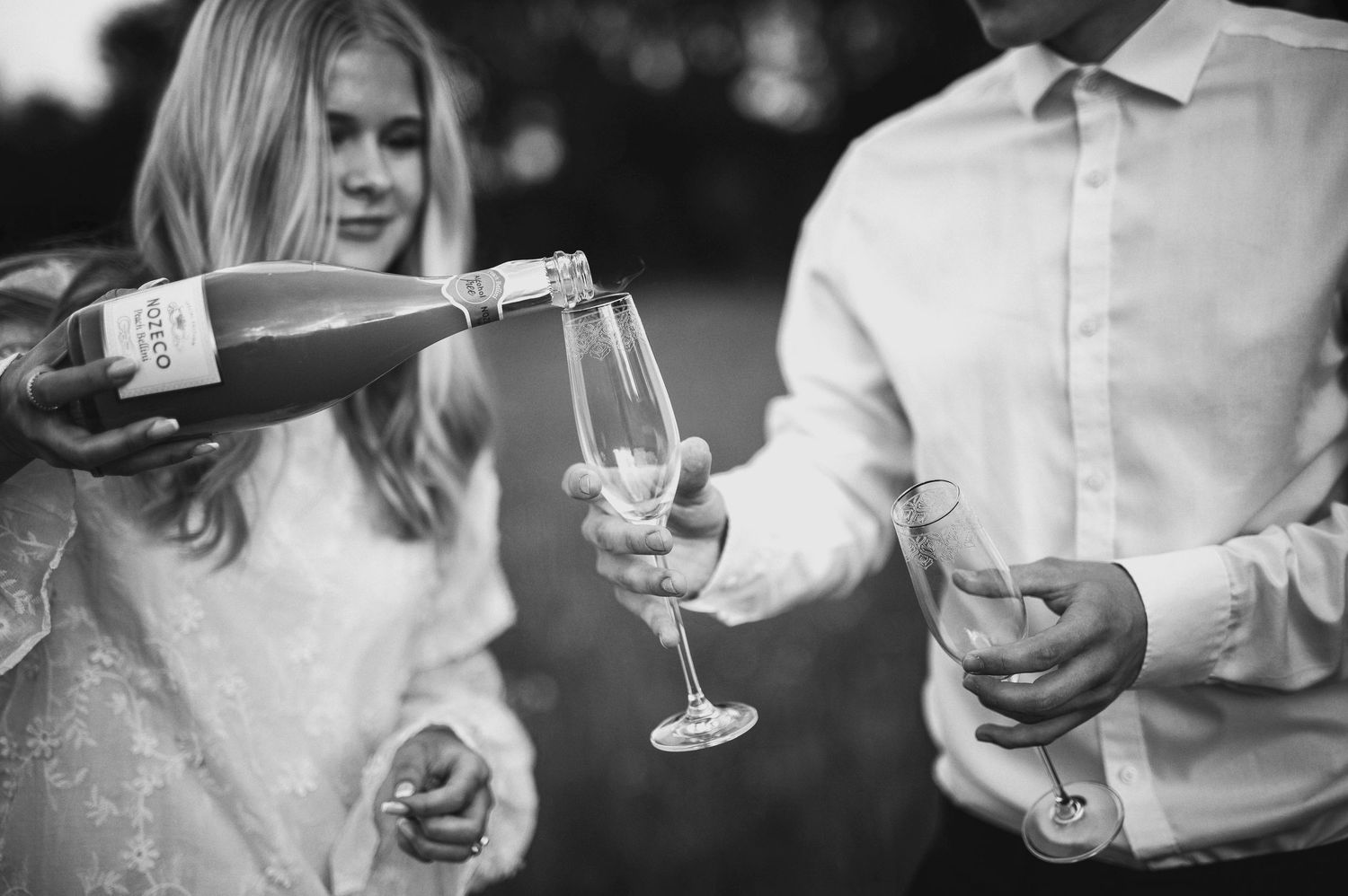 A black and white photo of champagne being poured into glasses held by a couple in elegant white attire.