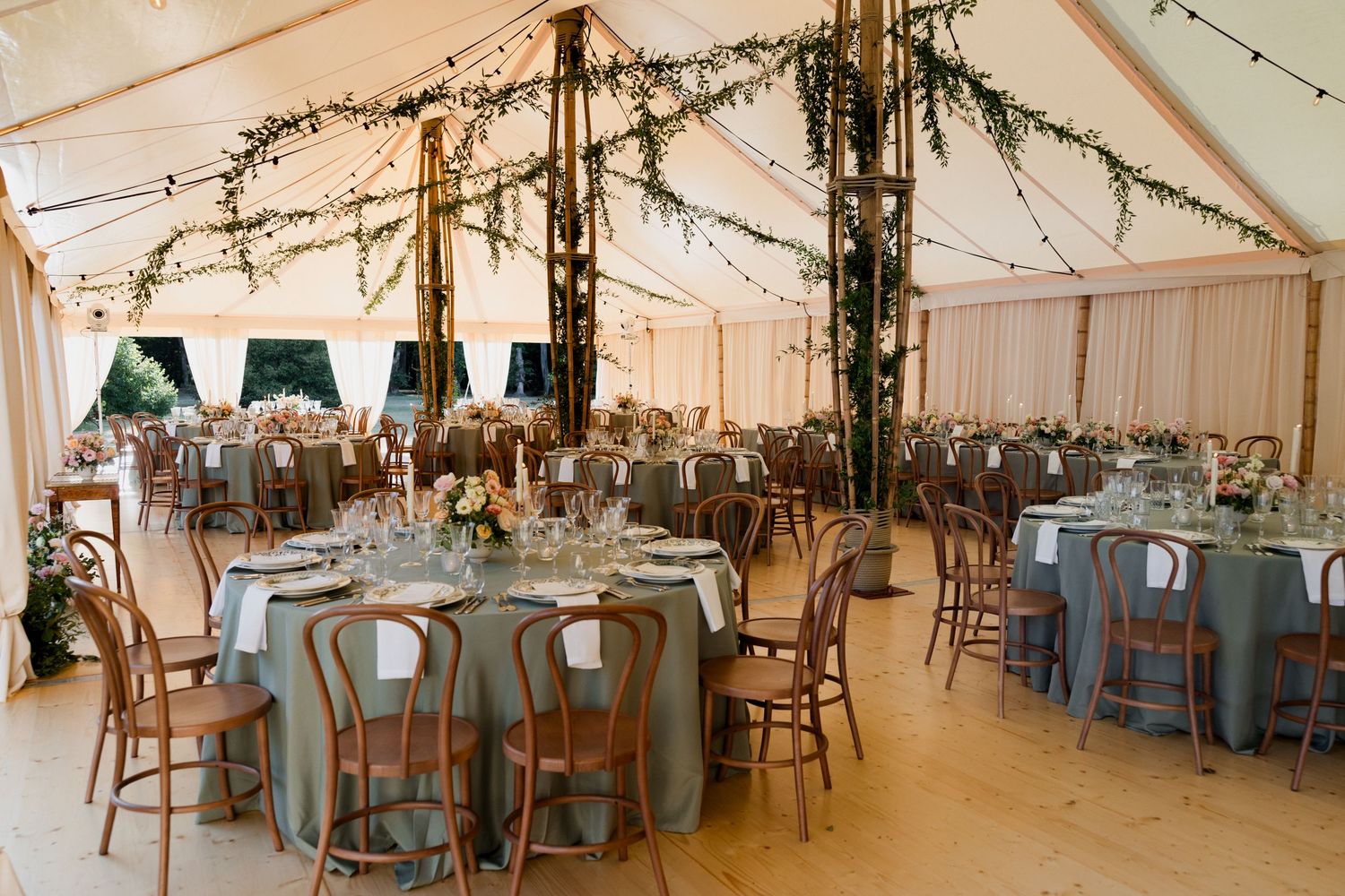 Wedding reception setup in an elegant white tent with round tables, blue linens, and twinkling string lights overhead.