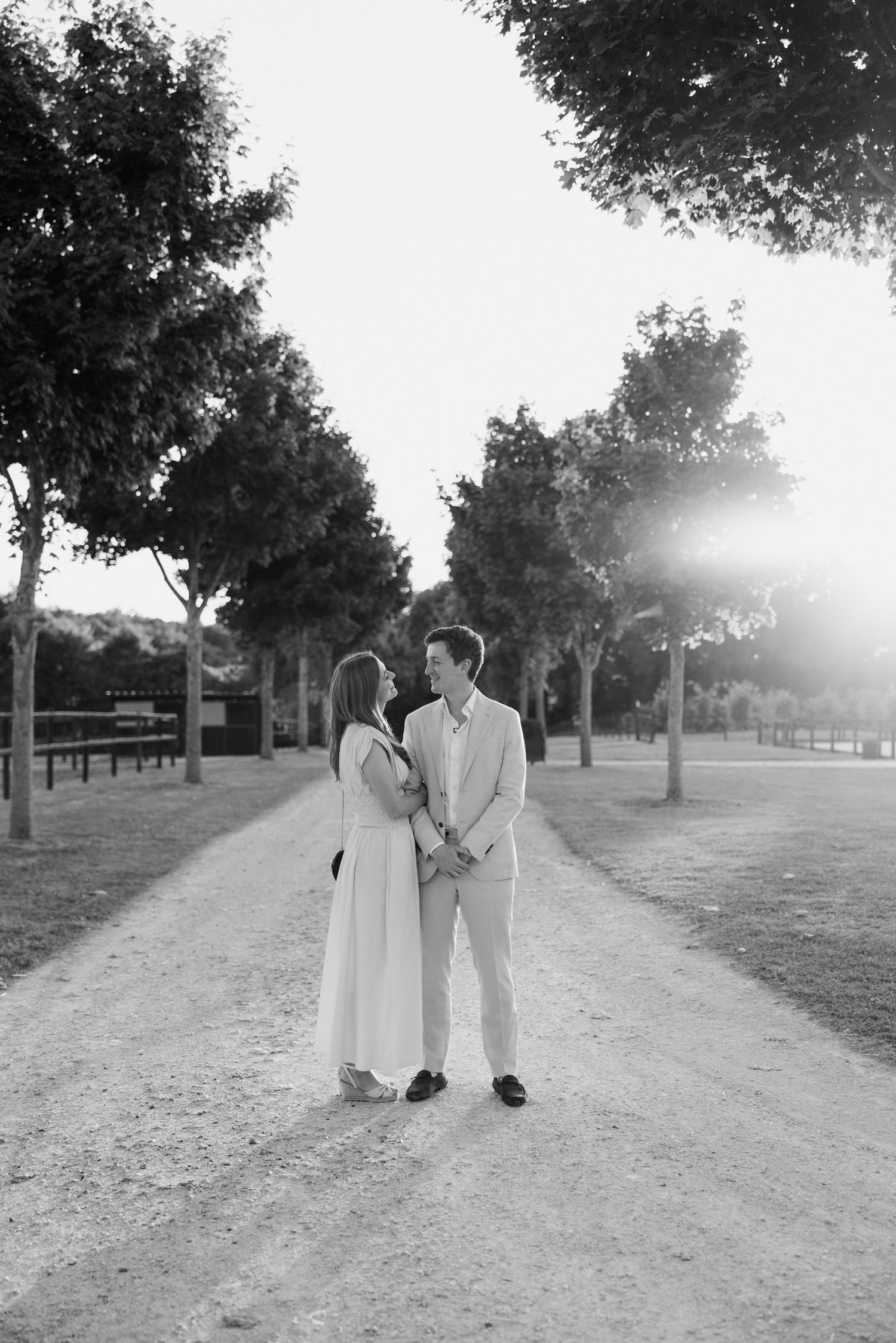A couple in white attire walks together on a tree-lined dirt path in a romantic outdoor setting.