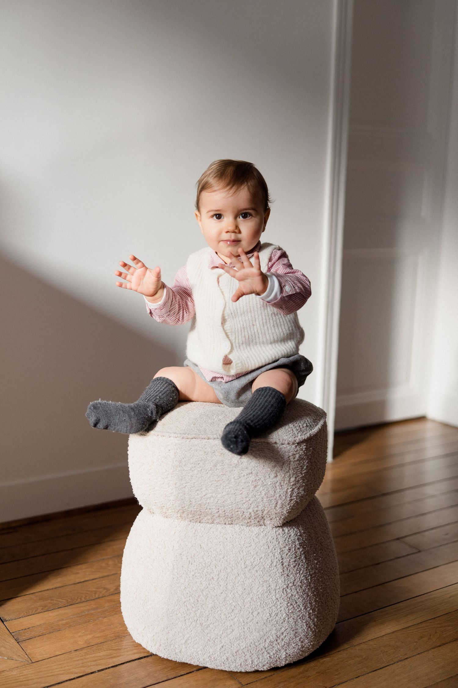 Cute baby sitting on a white boucle pouf stool, wearing a knit vest and gray socks, reaching out with both hands.