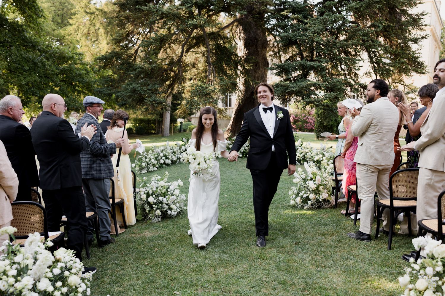 Green lawn outdoor wedding ceremony with guests gathering under tall trees at dusk.