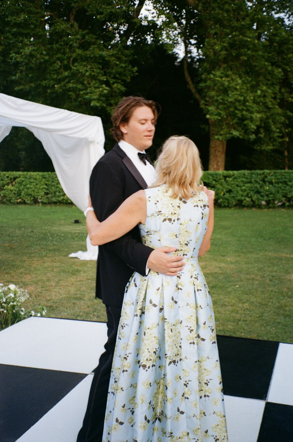 A couple shares a romantic dance together outdoors in formal attire, with the woman wearing a floral print dress.