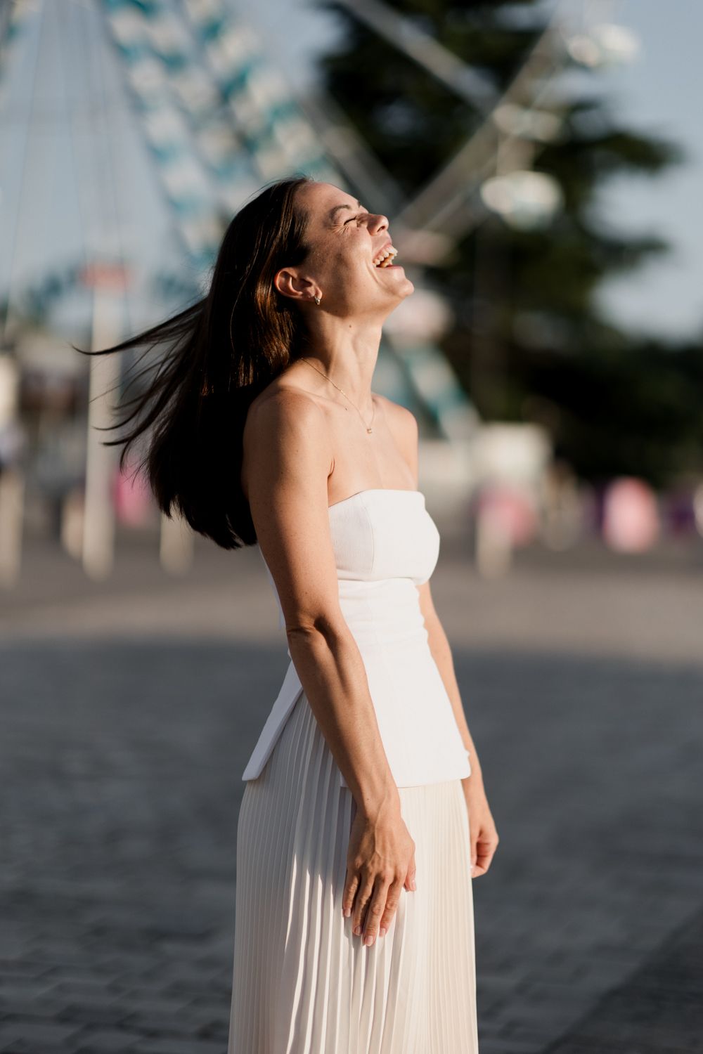 Woman in flowing white dress twirls joyfully on beach at sunset, hair and fabric moving in the wind.