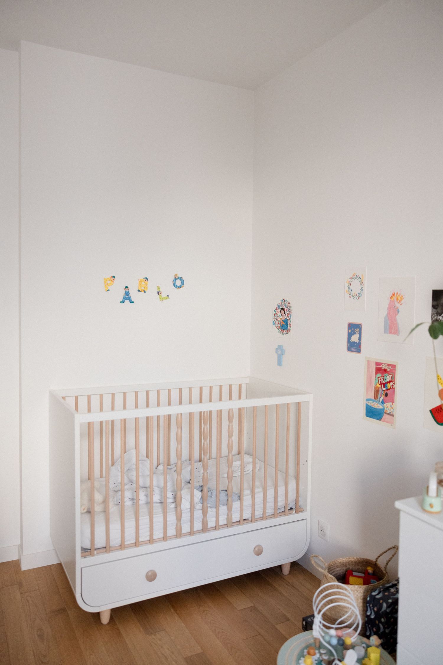 White modern baby crib in minimalist nursery with wooden floor and colorful wall decorations spelling a name.