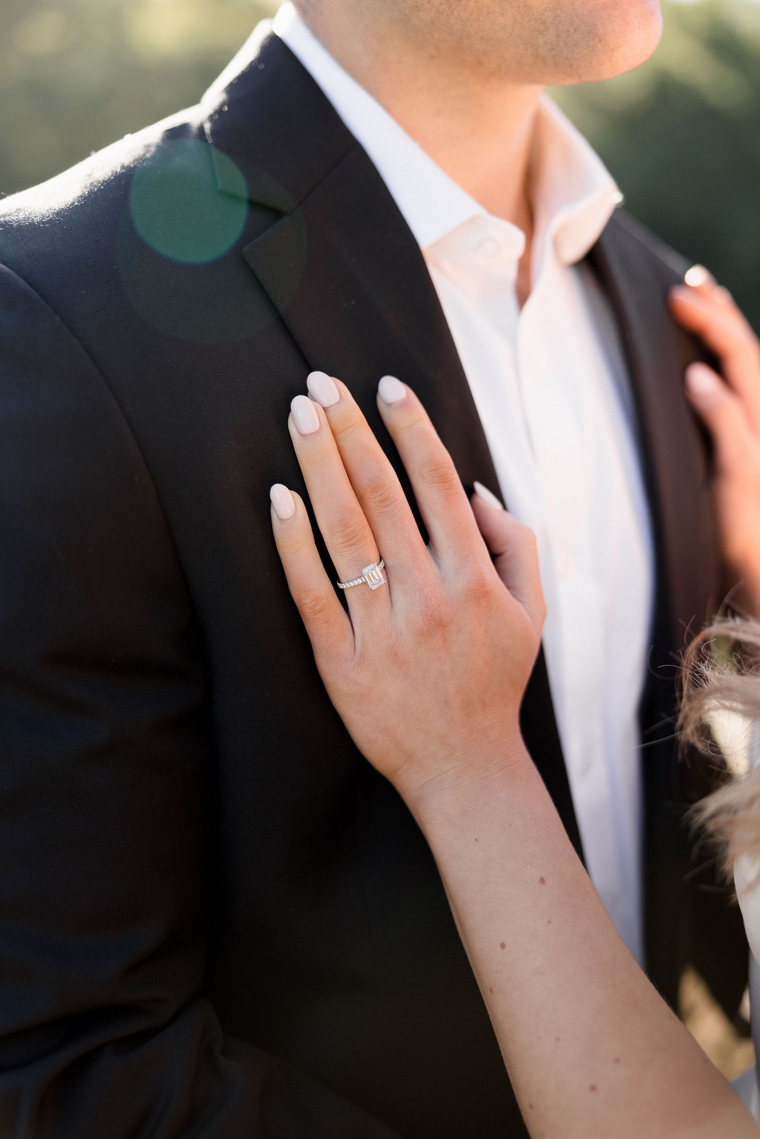 Bride's hand with diamond engagement ring and nude manicure resting on groom's black suit jacket.