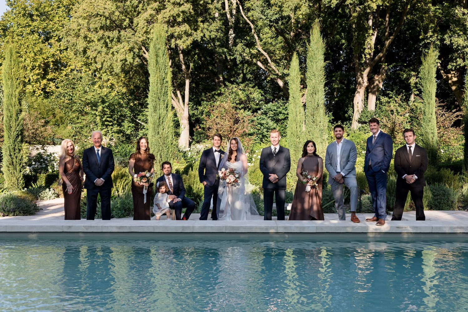 Wedding guests stand in line along a tranquil pond's edge surrounded by trees in a garden setting. wedding photographer france