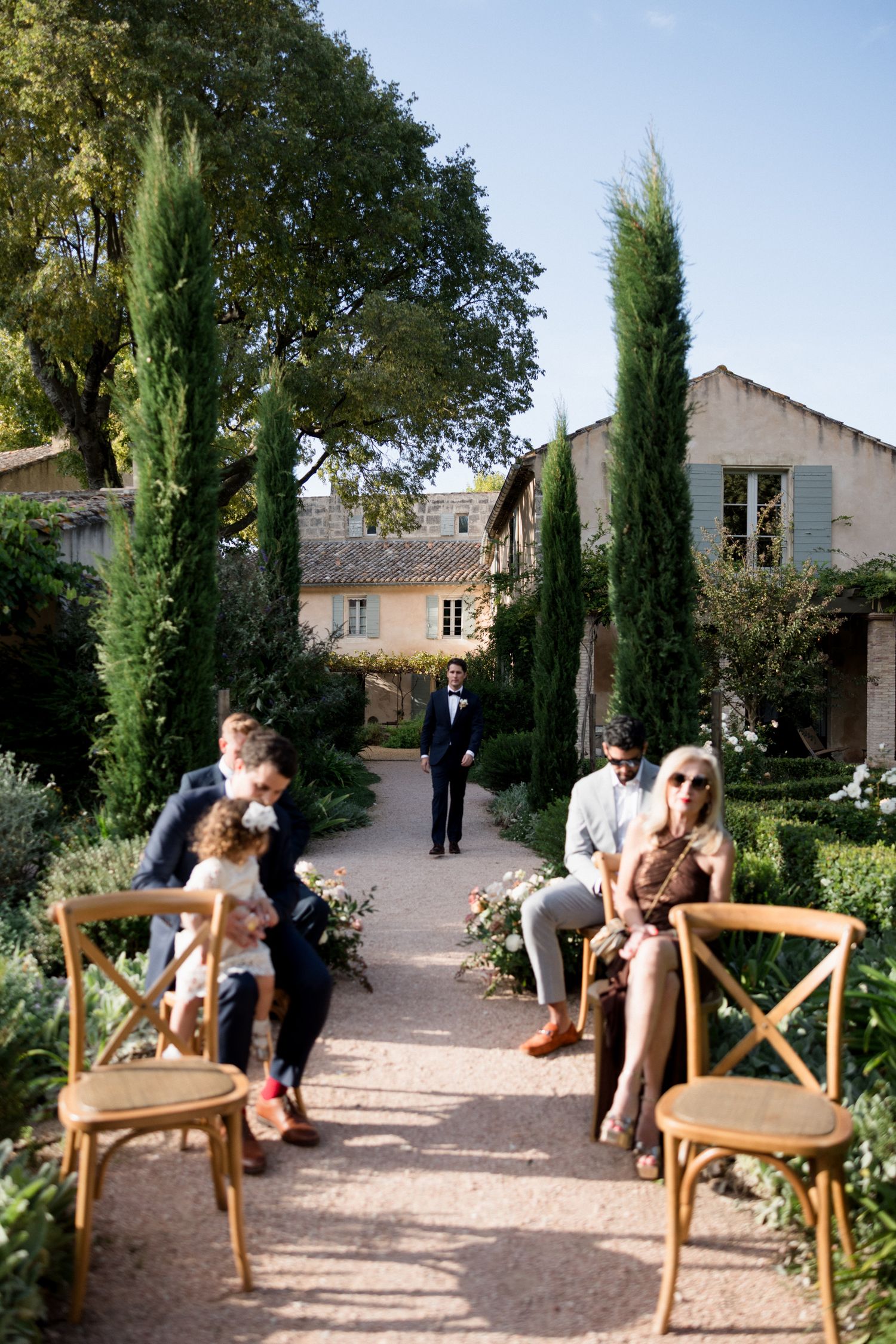 Wedding guests relax in wooden chairs along a garden path lined with cypress trees at an outdoor venue.