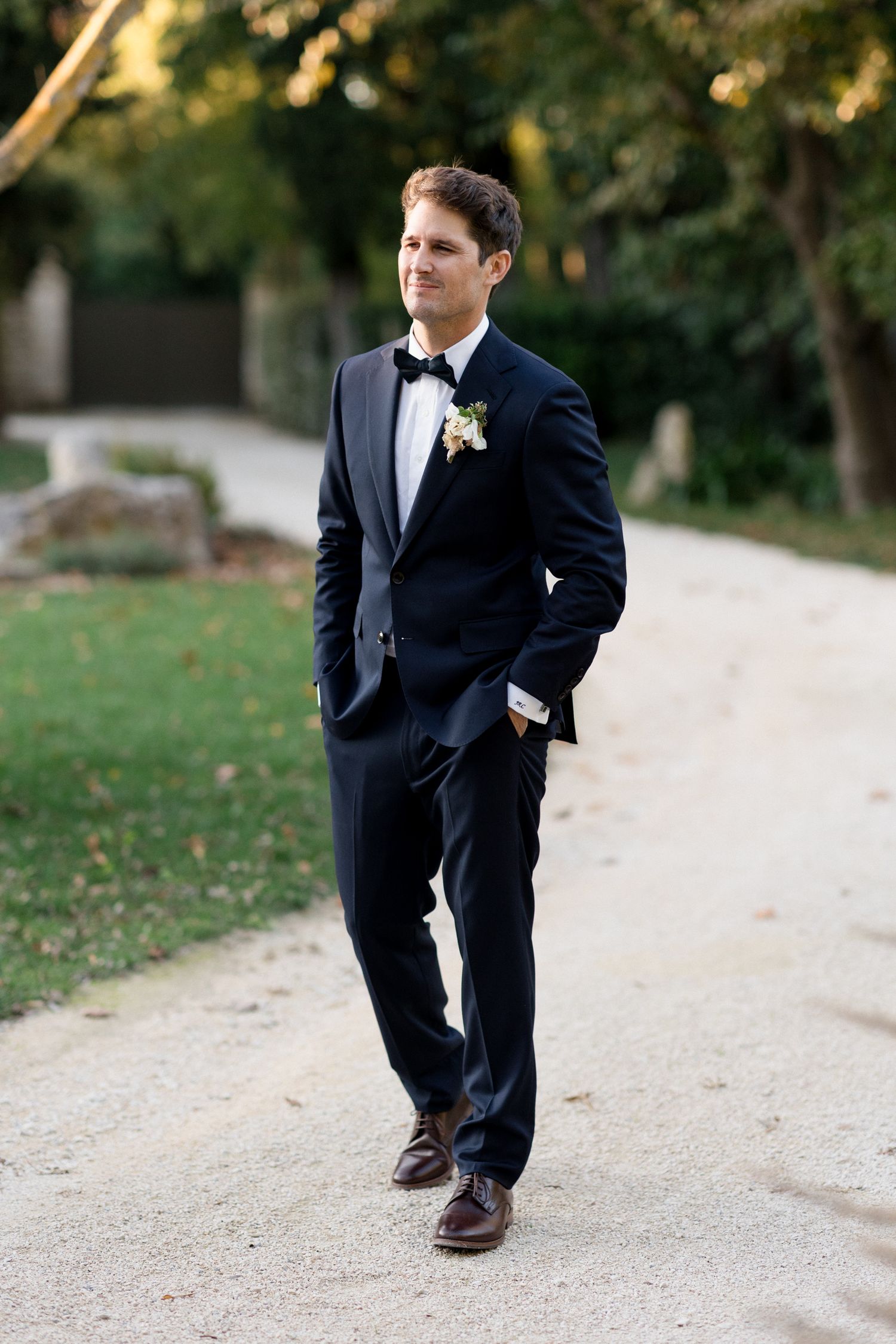 A person in a formal black tuxedo and bow tie poses outdoors on a path surrounded by autumn foliage.