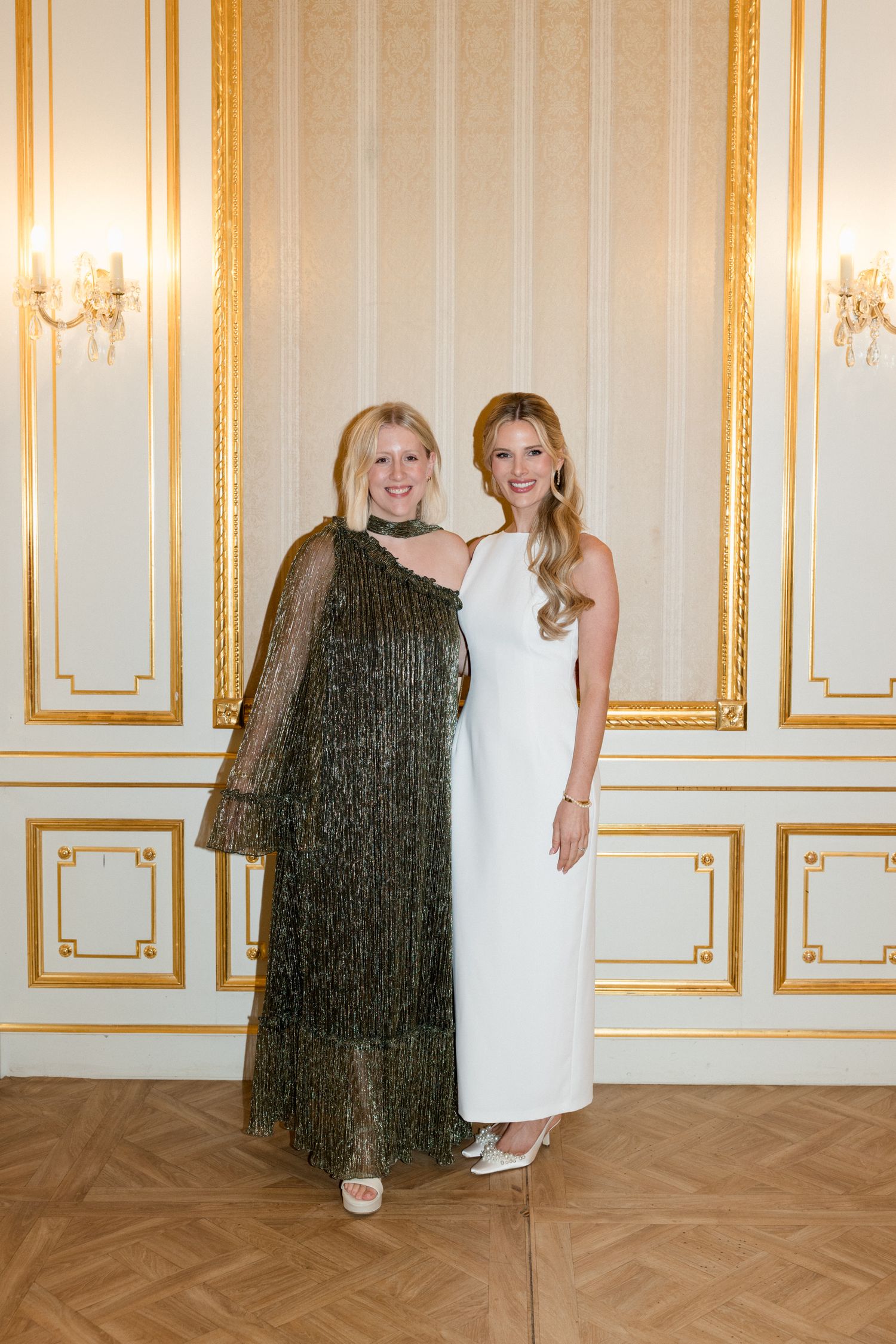 Two people in white dresses stand in an elegant room with gold-trimmed walls and herringbone wood floors.