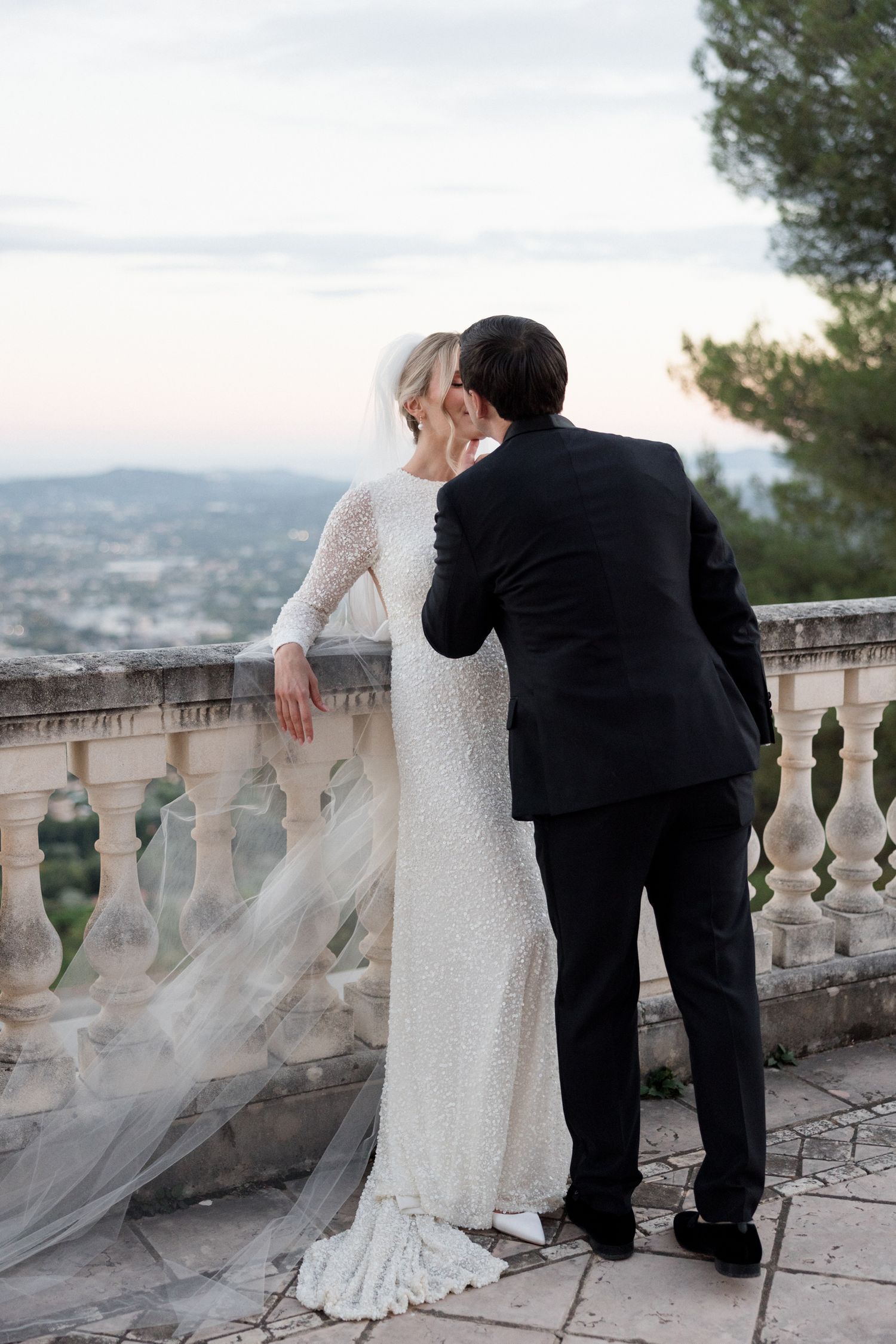 A couple in wedding attire embrace while looking out over a scenic mountain view from a stone balcony with ornate railings.