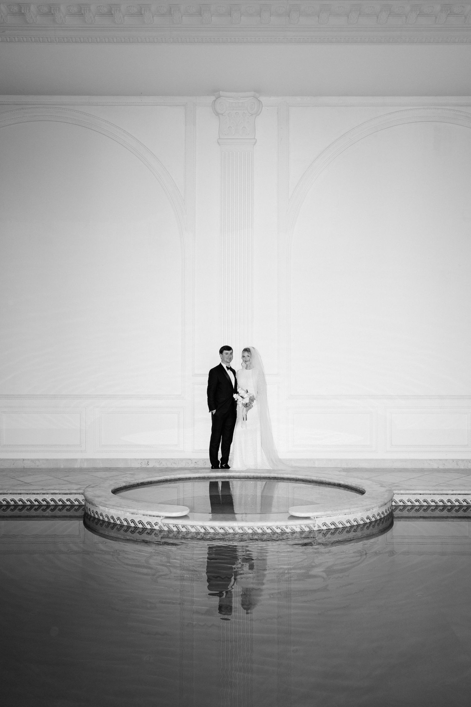 Black and white wedding photo of bride and groom standing on a circular platform reflected in water inside an elegant hall.