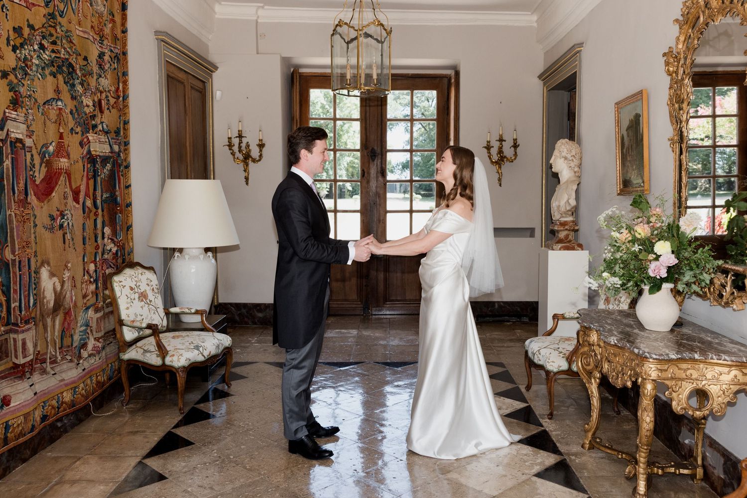 A couple shares their first look moment in an elegant room with ornate gold decor and large windows.