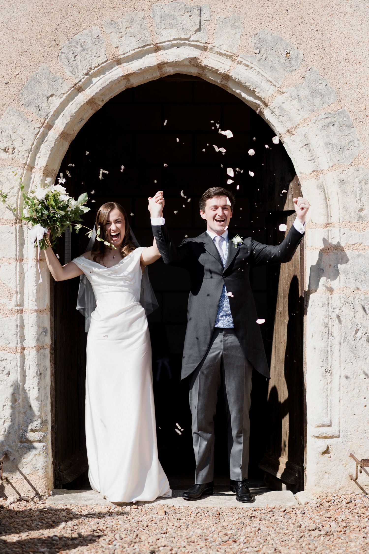 Newlyweds celebrate with joy under a stone archway entrance as confetti falls around them at their wedding ceremony. Paris wedding photograp