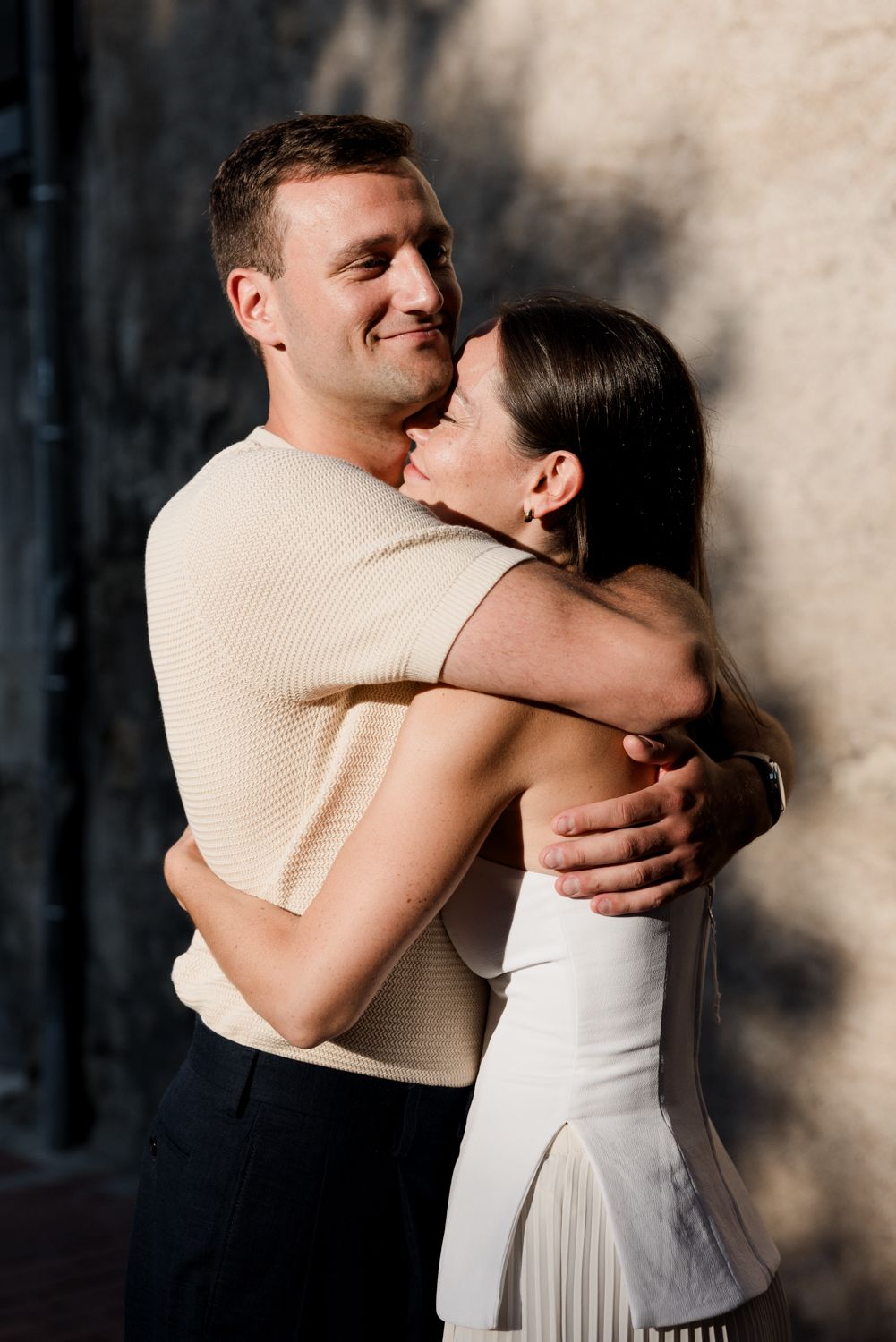 A romantic photo sequence showing a couple in a gentle embrace against a shadowed wall, wearing cream and white clothing. Engagement session