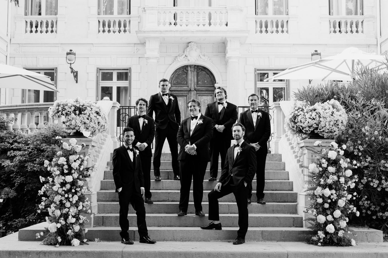 Five formal outdoor portraits showing groups wearing black tuxedos on steps with white floral arrangements on a sunny day.