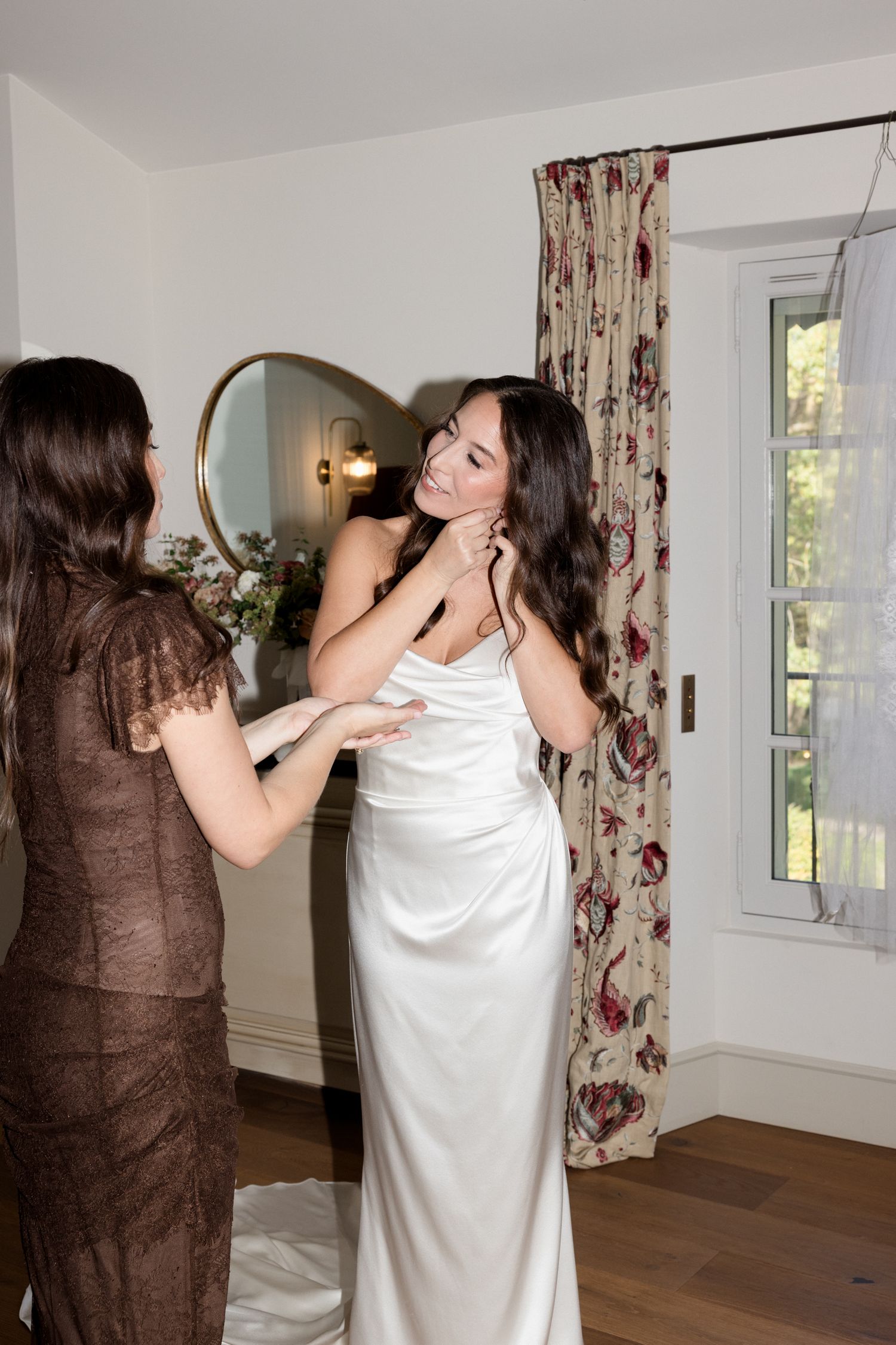 Two people in white and brown attire interact playfully in a bright room with floral curtains and hardwood floors. Editorial wedding photogr
