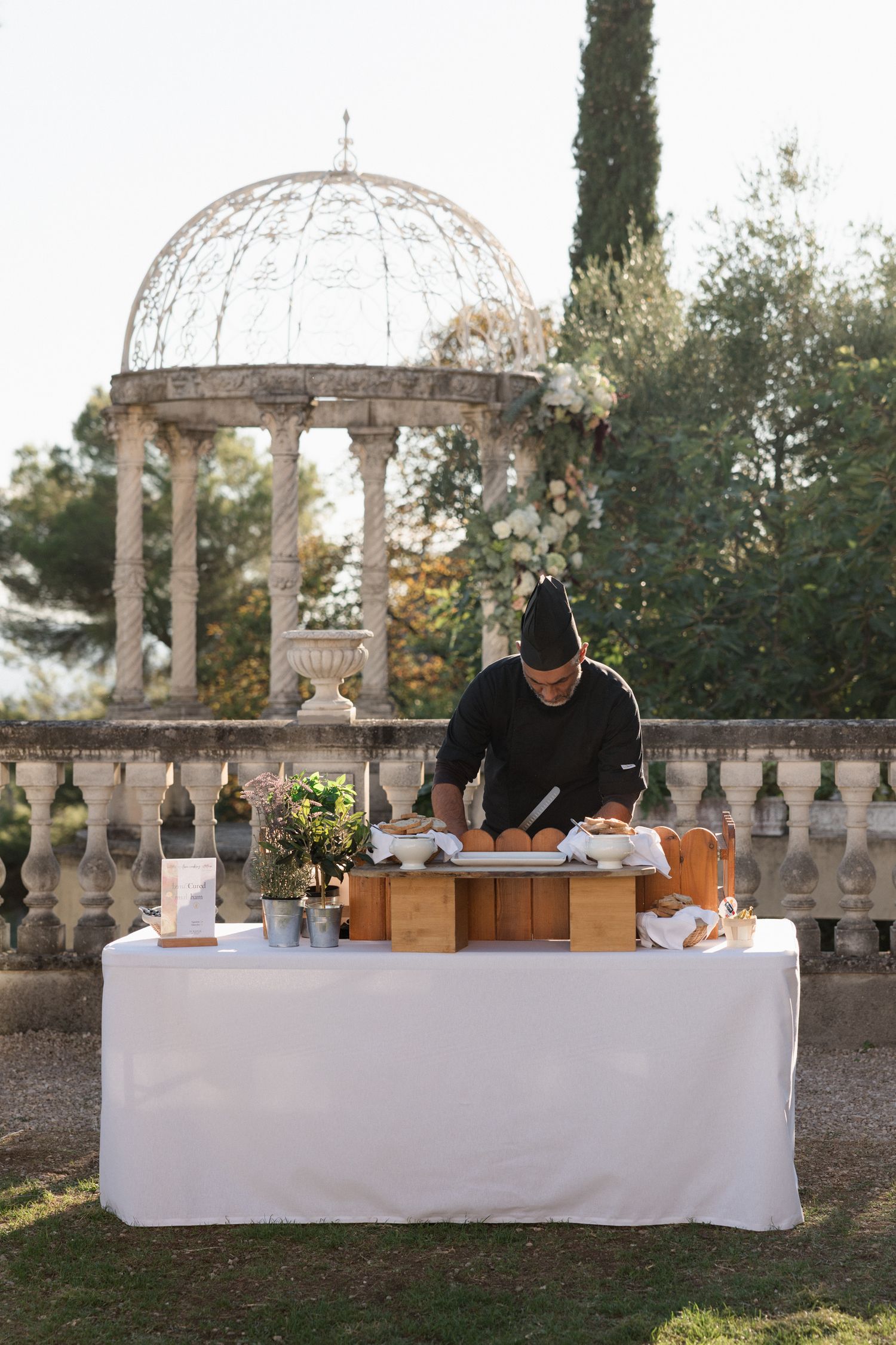 A person arranges items on a white-clothed table against an ornate garden backdrop with cypress trees and a gazebo.