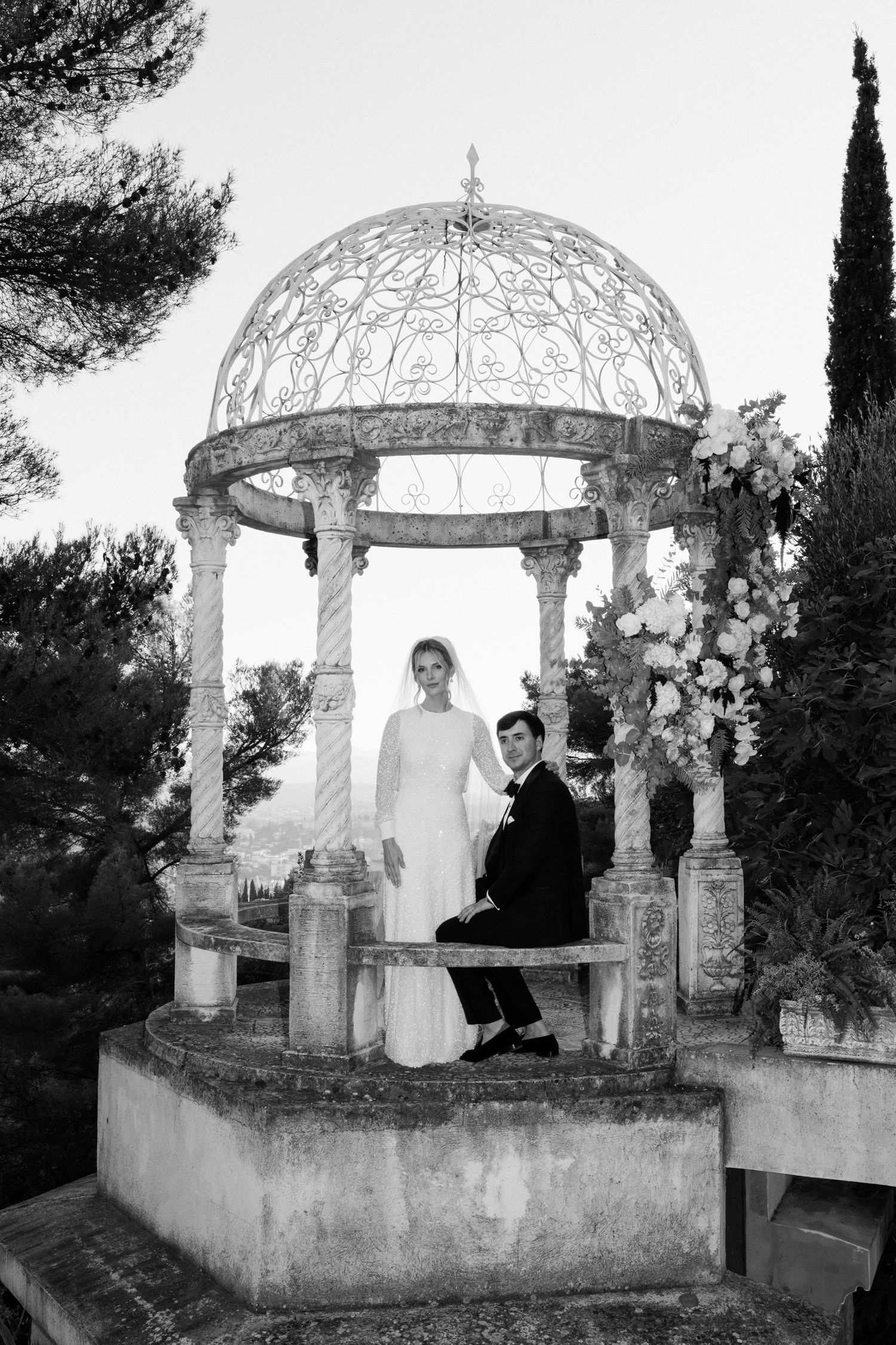 A black and white photograph of a couple sitting in an ornate stone gazebo with a domed roof and decorative columns.