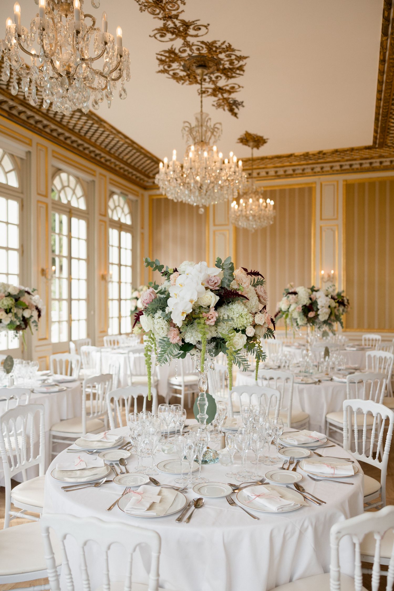 Round dining tables with white linens and floral arrangements set beneath ornate chandeliers in a classic ballroom setting. destination wedd