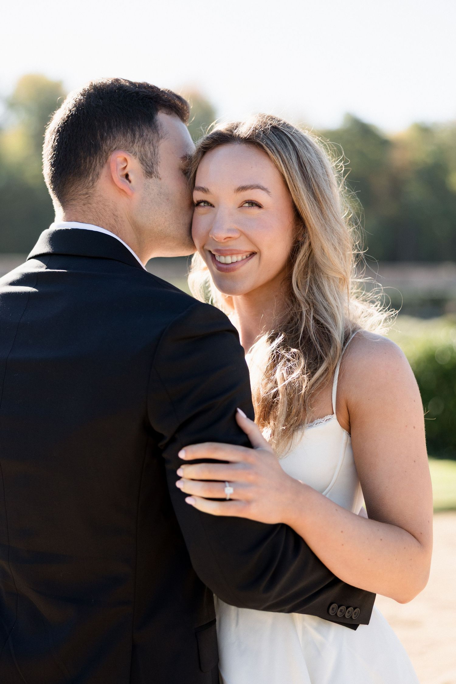 Smiling bride in a white dress embracing groom in black suit outdoors in golden sunlight, showing engagement ring.