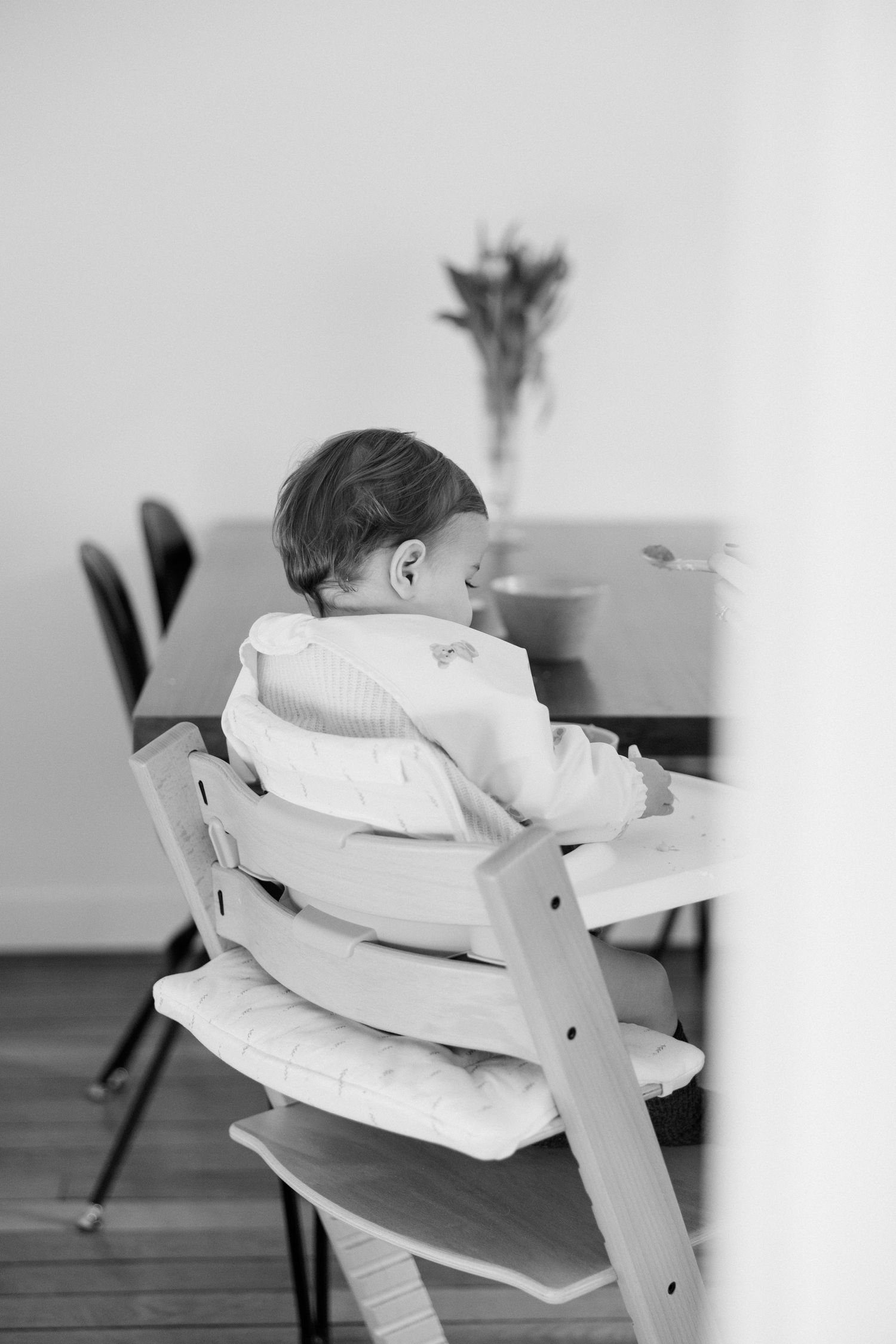 Black and white photo of a baby sitting in a Stokke Tripp Trapp high chair at a dining table.