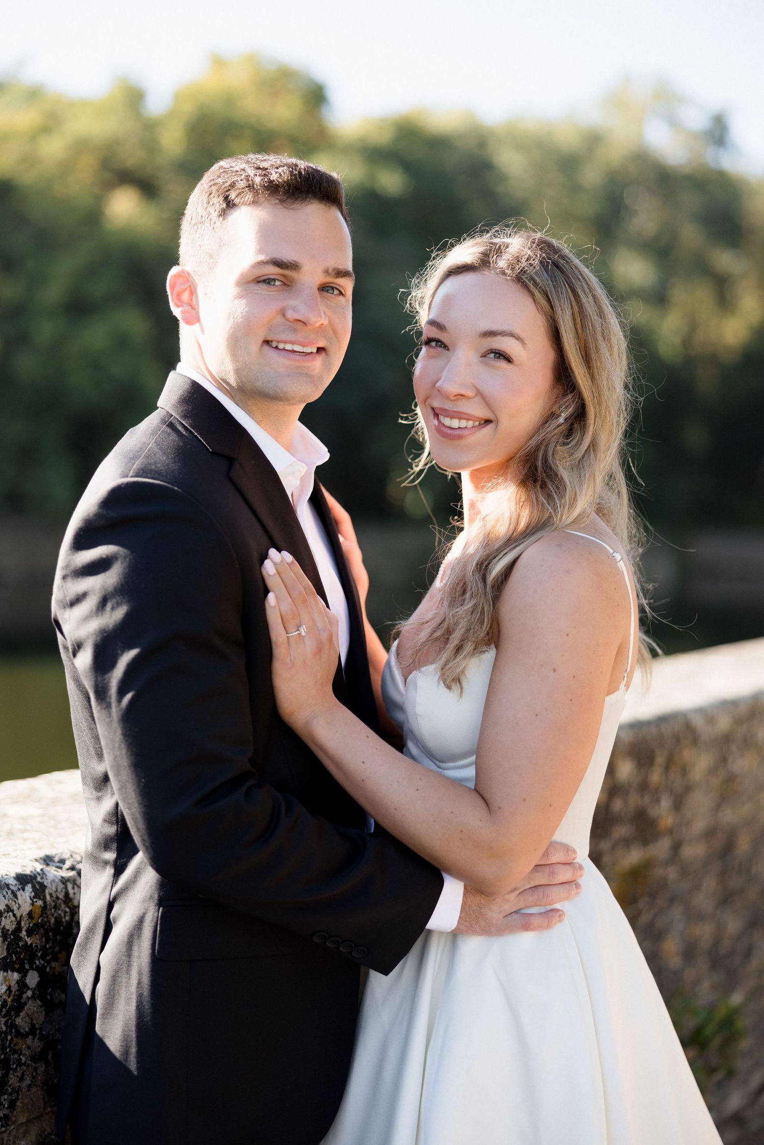 Smiling bride and groom embrace outdoors near a sunlit waterway, wearing wedding attire.