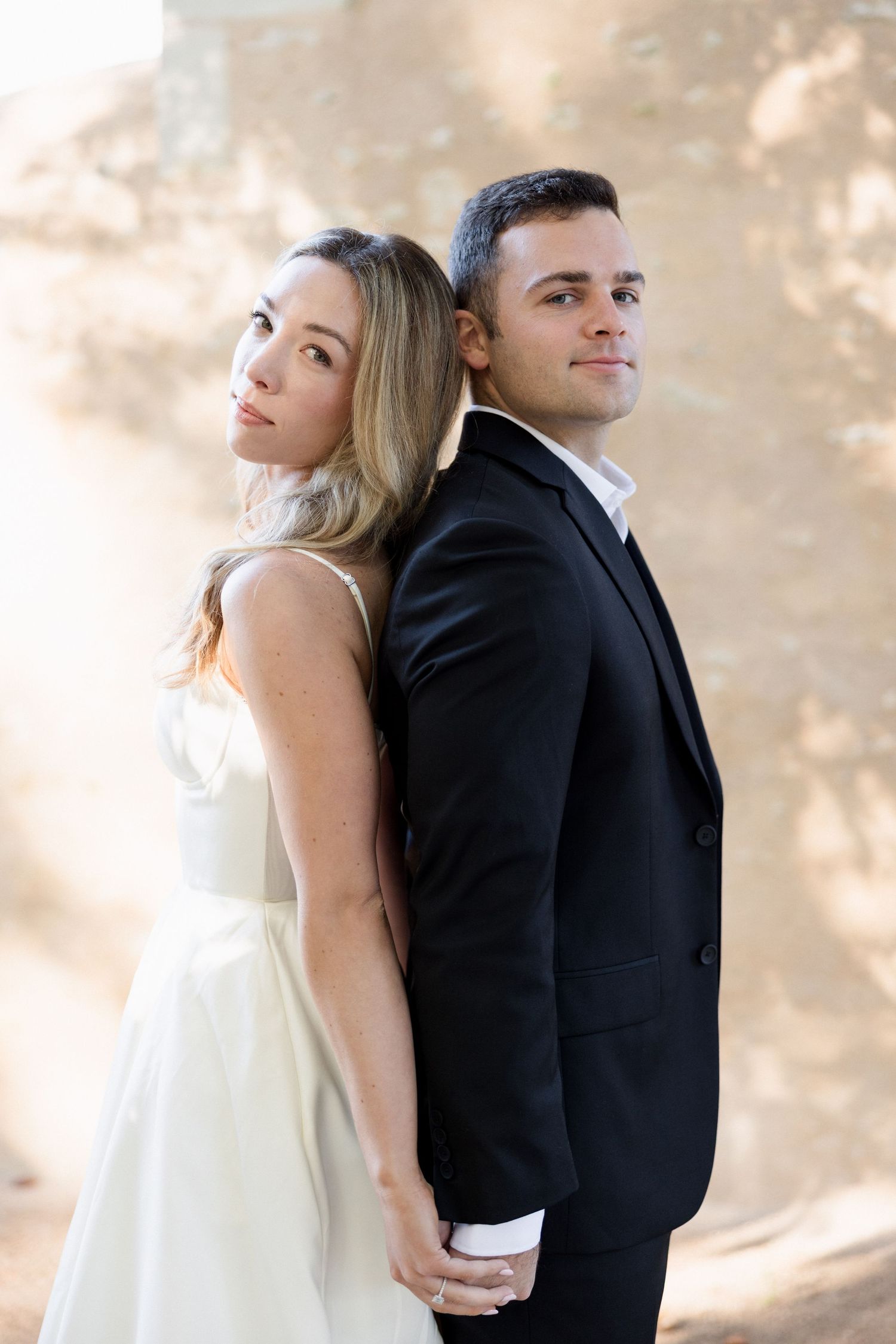 Elegant couple in wedding attire posing back-to-back, bride in white gown holding grooms hand outdoors.