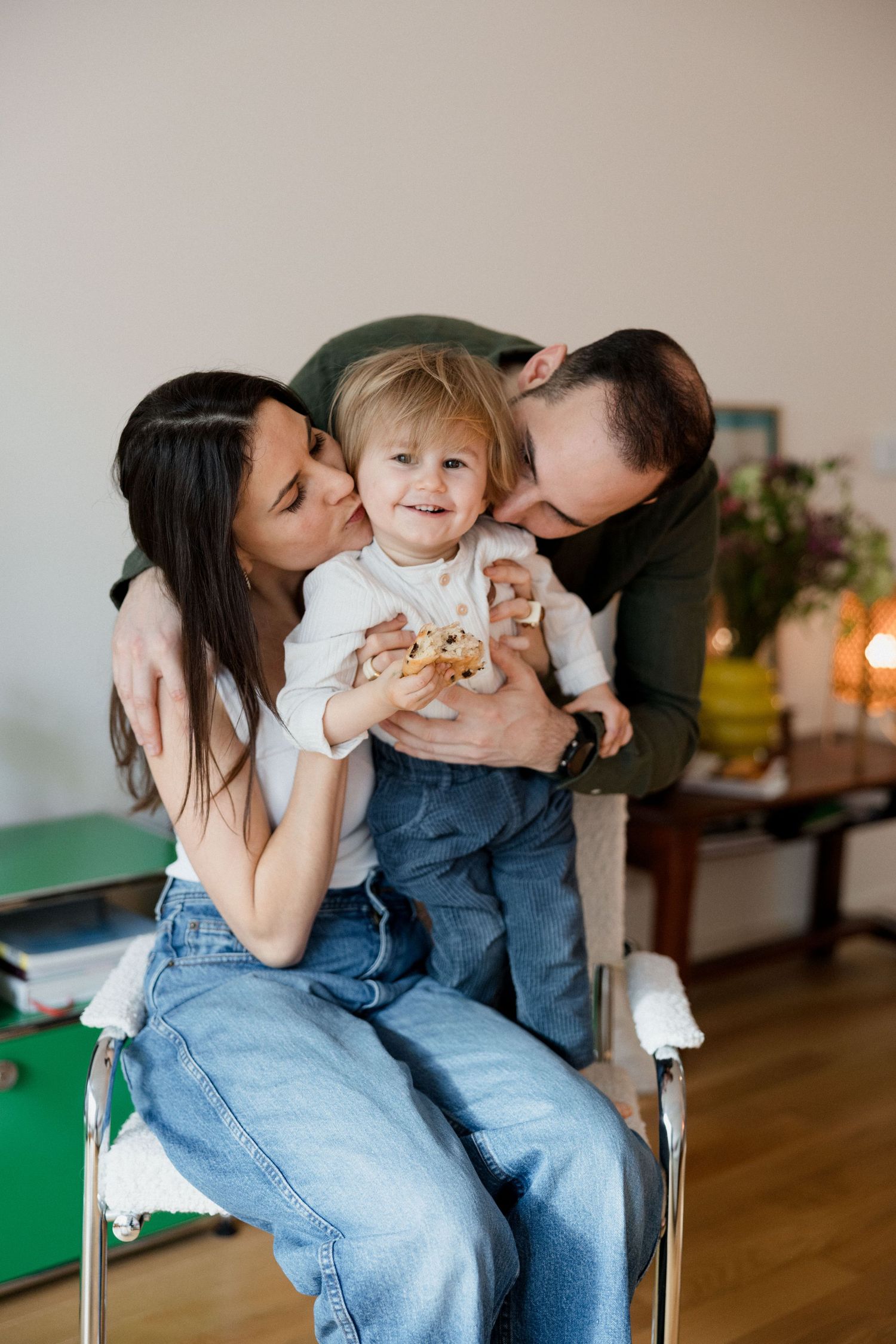 Happy family moment with parents kissing their smiling toddler holding a cookie while sitting on a chair indoors.